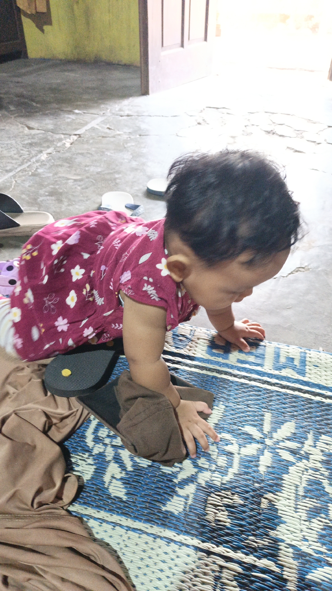 In the city of Sambi, Indonesia, a toddler in a maroon dress with a floral pattern crawls on a woven mat. The mat, featuring a blue and white floral design, is laid on a cracked concrete floor. The toddler's hands are on the mat, and a brown cloth lies to their left. To the left of the mat, a dark-colored sandal is visible, and further left, other light-colored footwear can be seen on the concrete floor. The background shows a doorway with a wooden door and a wall painted yellow. Sunlight illuminates the scene from the right, casting shadows.
