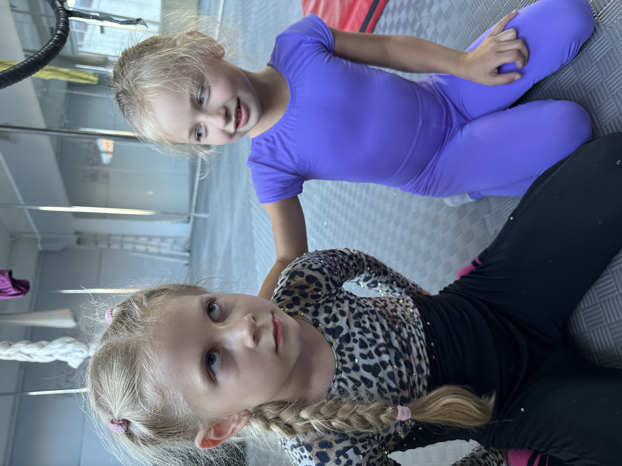Two young girls are sitting on a padded floor, likely in a gymnastics or dance studio. The girl in the foreground has blonde hair in a braid with a pink elastic and is wearing a black leotard with a leopard print overlay and black leggings with pink stripes. The girl in the background is wearing a purple leotard. Both girls appear to be of Caucasian ethnicity and are looking in different directions. The lighting suggests it is daytime. There is no visible text in the image.