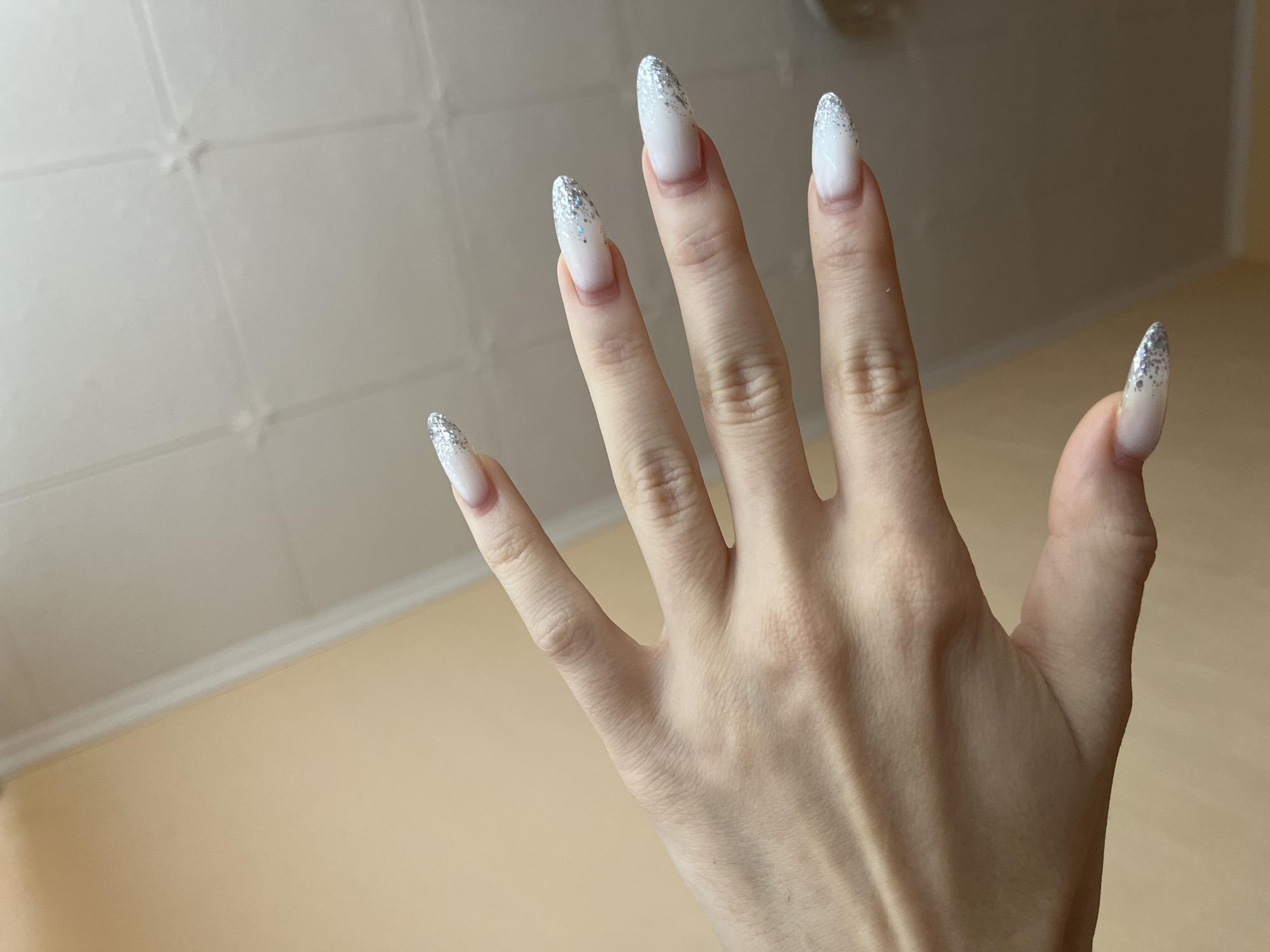 A close-up, top-down shot shows a person's left hand and forearm against a neutral background. The hand is held palm-up, fingers slightly splayed, with nails painted white with a glittery silver gradient effect. The skin appears smooth with visible veins. The background is divided into two areas: a light peachy-toned wall on the left and a textured, off-white ceiling with a grid pattern on the right. The image does not contain any information that can be used to determine its location within the city of Игнатово, Russia.