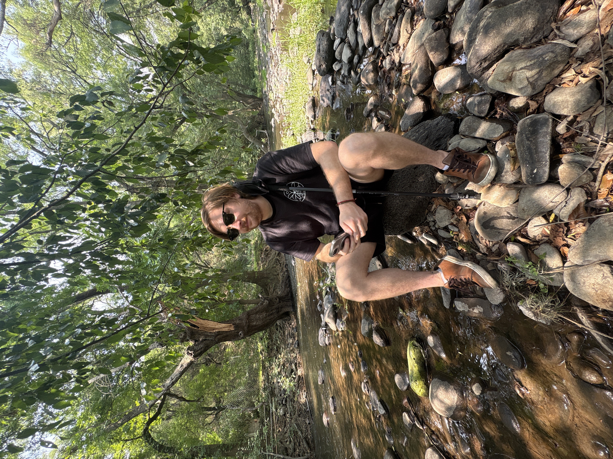 A man is sitting on a rock in a creek. He is wearing sunglasses, a black t-shirt, and brown boots. The creek is lined with rocks and trees. The location is Cañada de Madero, Mexico.