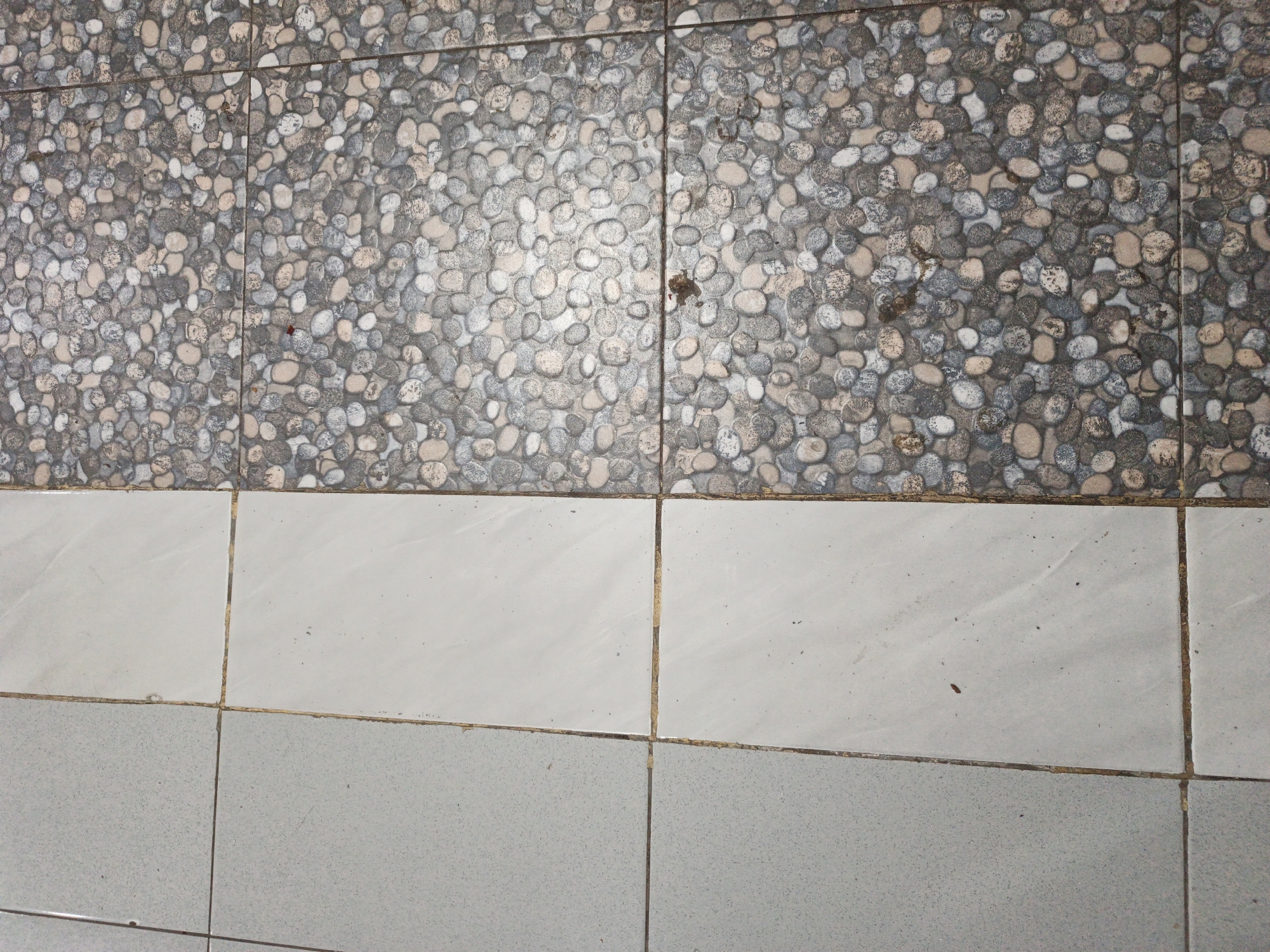 The image displays a close-up, overhead view of a tiled surface in Kelapapati, Indonesia. The upper portion of the image features tiles with a mosaic pattern of small, rounded stones in various shades of gray, beige, and white. This mosaic tile section transitions horizontally to a band of larger, square tiles. The top band of these larger tiles is a solid, light cream color, while the lower band is a solid light gray. The grout lines between all the tiles are visible and appear discolored with a yellowish-brown hue. The surface appears to be outdoors, possibly a walkway or patio, given the texture and the potential for weathering. There are no people or discernable actions depicted in the image.