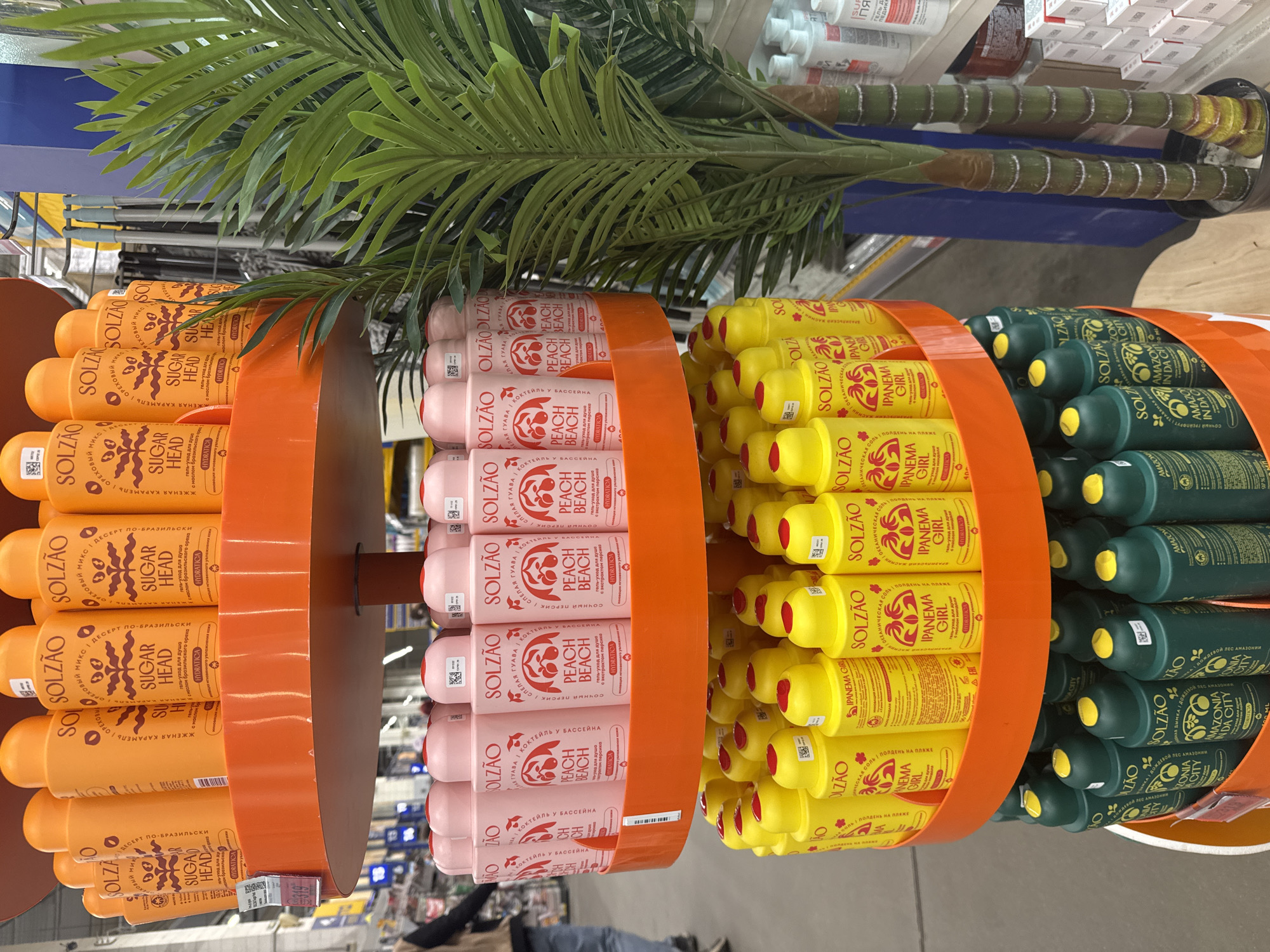 The image shows a retail display featuring Solzao brand personal care products, likely shower gels, organized by scent and color. The display has orange circular frames holding rows of bottles. The scents, indicated on the labels, include "Sugar Head" (orange bottles), "Peach Beach" (pink bottles), "Ipanema Girl" (yellow bottles), and "Amazonia" (green bottles). An artificial palm tree is positioned above the display. The image was likely taken inside a retail store. No specific details indicate that this image comes from Saint Petersburg, Russia. There is no visible information available regarding actions, interactions, and the environment.