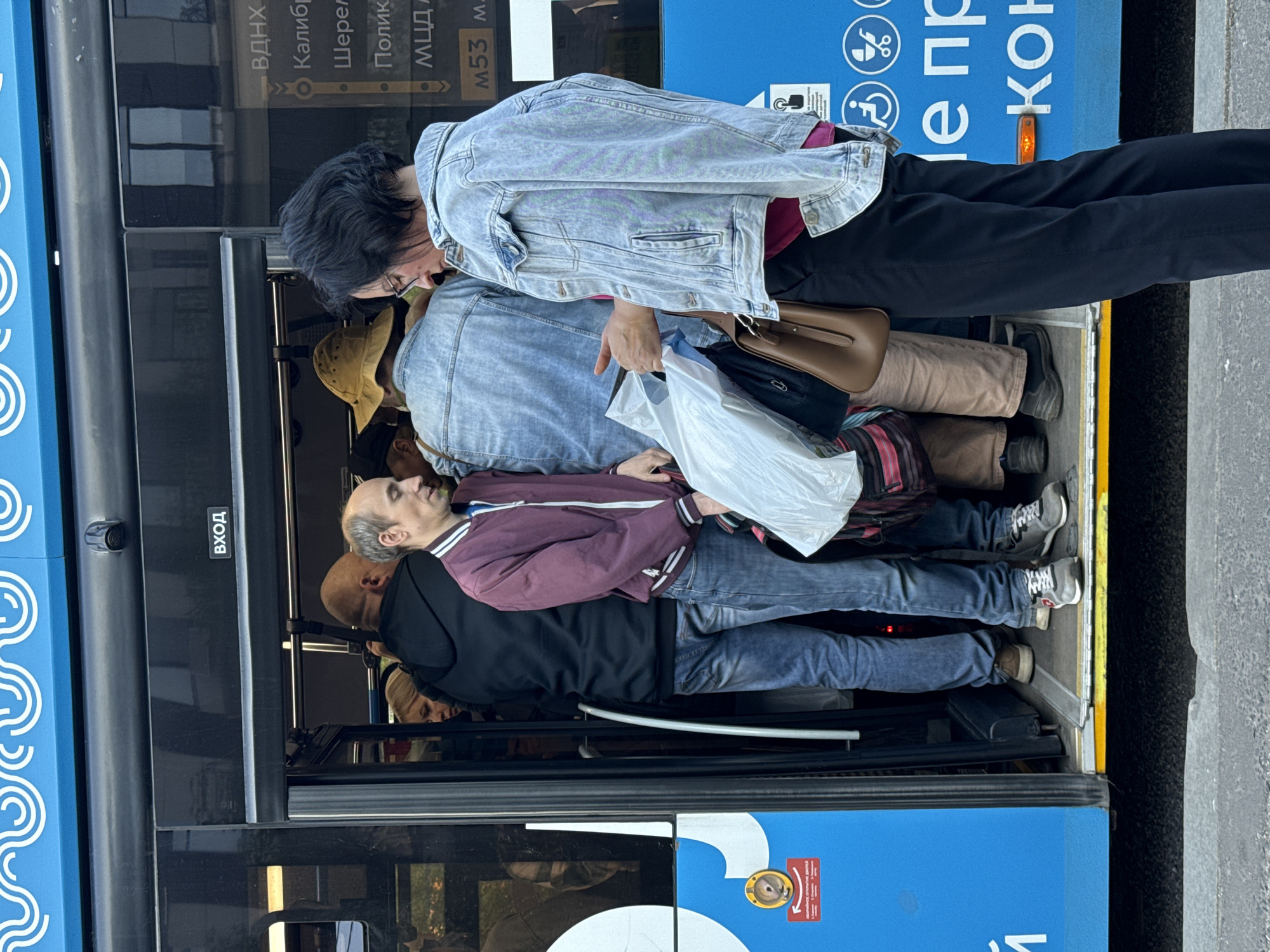 The photograph shows a crowded bus in Moscow, Russia, with passengers packed tightly together at the entrance. The scene is captured from an external perspective, looking into the bus through the open doors.
Several people are visible in the frame, standing shoulder-to-shoulder. Some passengers are wearing casual attire, including jeans, jackets, and shirts. One individual holds a white plastic bag. Others carry purses or bags.
The interior of the bus is visible, and there are handles and rails for passengers to hold onto. The exterior of the bus has blue accents with white swirl designs. Russian text is visible on the side of the bus, providing destination information.
The scene indicates a typical urban environment during rush hour or a period of high transit demand.