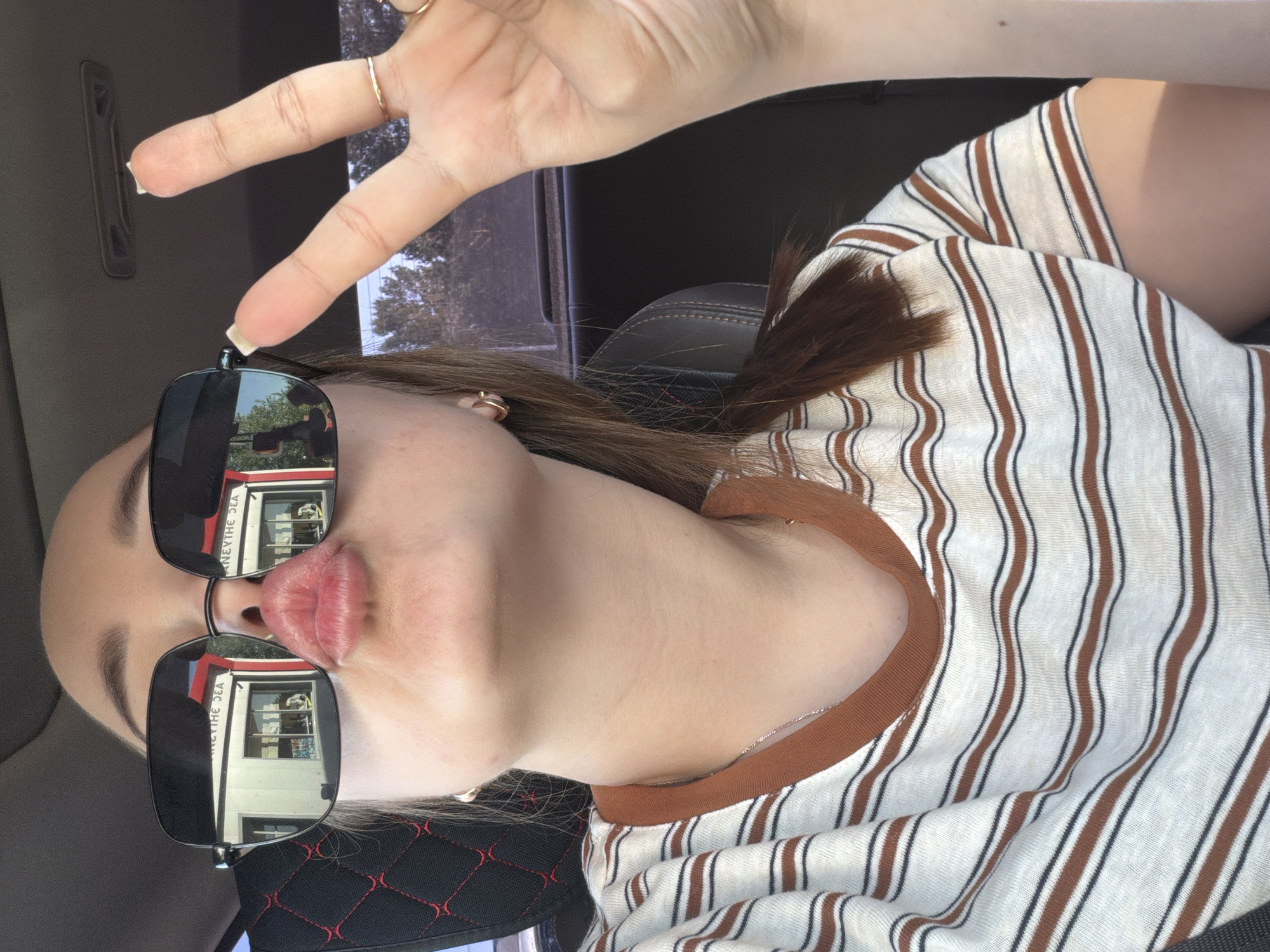 A selfie of a person wearing sunglasses in the passenger seat of a car. The reflection in the sunglasses shows a building labeled "ANEXTHE SEA." This business is located in the city of Chaikovsky, Russia. The person is making a peace sign. They are wearing a striped shirt.