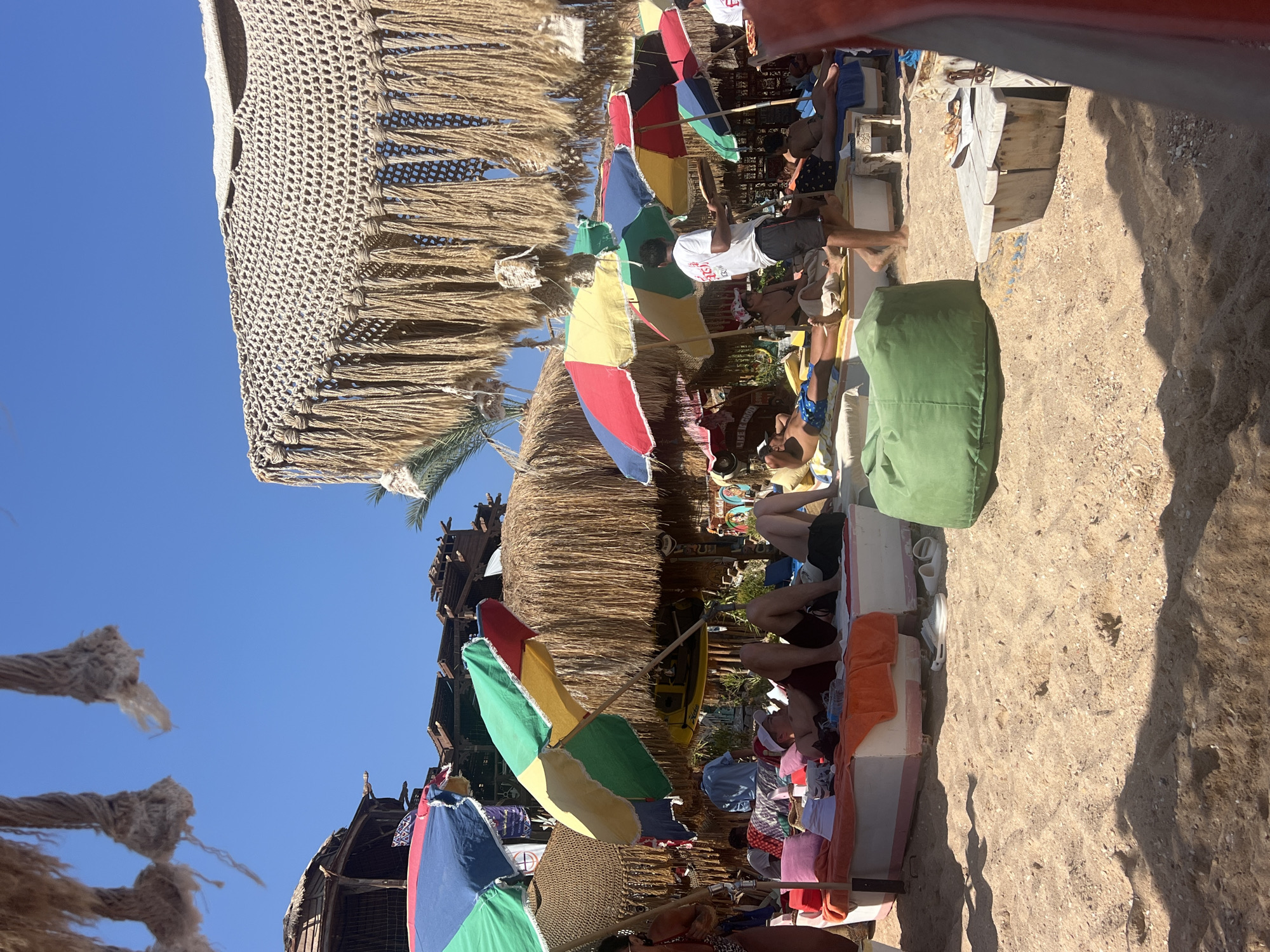 This image depicts a lively beach scene at a resort in Sharm El Sheikh, Egypt, under a clear blue sky. The foreground is dominated by sandy ground, with some shells and small pebbles visible. In the mid-ground, a raised, white rectangular platform is covered with orange, pink, and white cushions, serving as a lounging area for several individuals.
People are seen relaxing on this platform and on the sand. One person is lying down wearing a red hat and dark shorts, another is wearing a white t-shirt and dark shorts, and several others are partially visible, some in swimwear. A waiter in a white t-shirt and dark shorts is walking, carrying a tray with food. Various colorful umbrellas, including red, yellow, green, and blue ones, provide shade for the beachgoers.
Behind the lounging area, there is a thatched-roof structure and other rustic wooden buildings, suggesting resort infrastructure. A large, macrame-covered structure with fringes hangs from above, acting as a decorative canopy. Green beanbag chairs are scattered on the sand. The overall atmosphere appears relaxed and summery, indicative of a typical beach resort environment in Sharm El Sheikh.