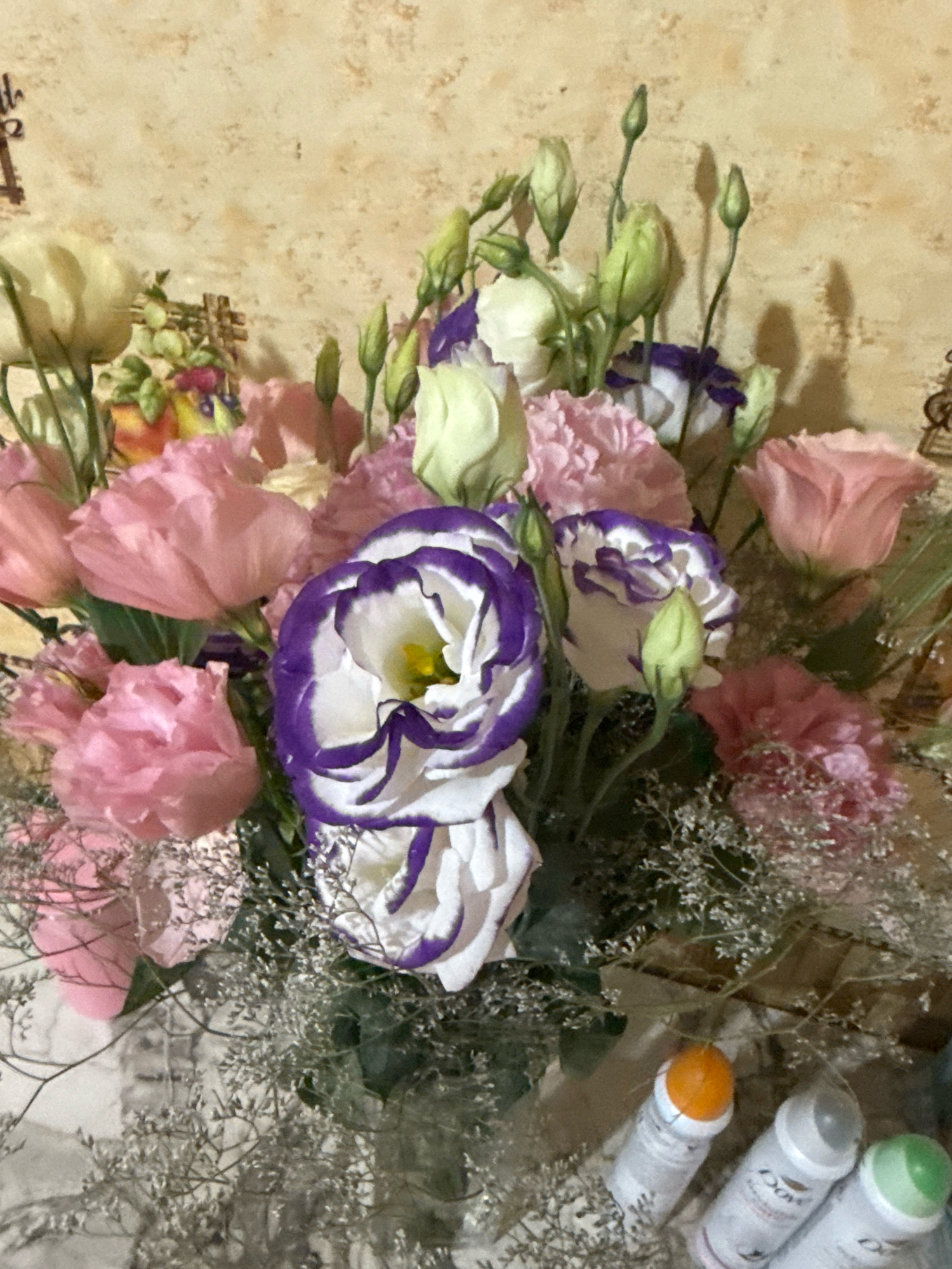 The image shows a bouquet of flowers against a textured, light-colored wall. The bouquet features pink carnations, white and purple lisianthus, and several green flower buds. There are also delicate dried flowers or filler in shades of gray and white. In the lower right corner, three roll-on deodorants are visible. The overall impression is that of a floral arrangement displayed indoors. The image does not contain any elements that would indicate its location in Basarabeasca, Moldova, or any specific city.