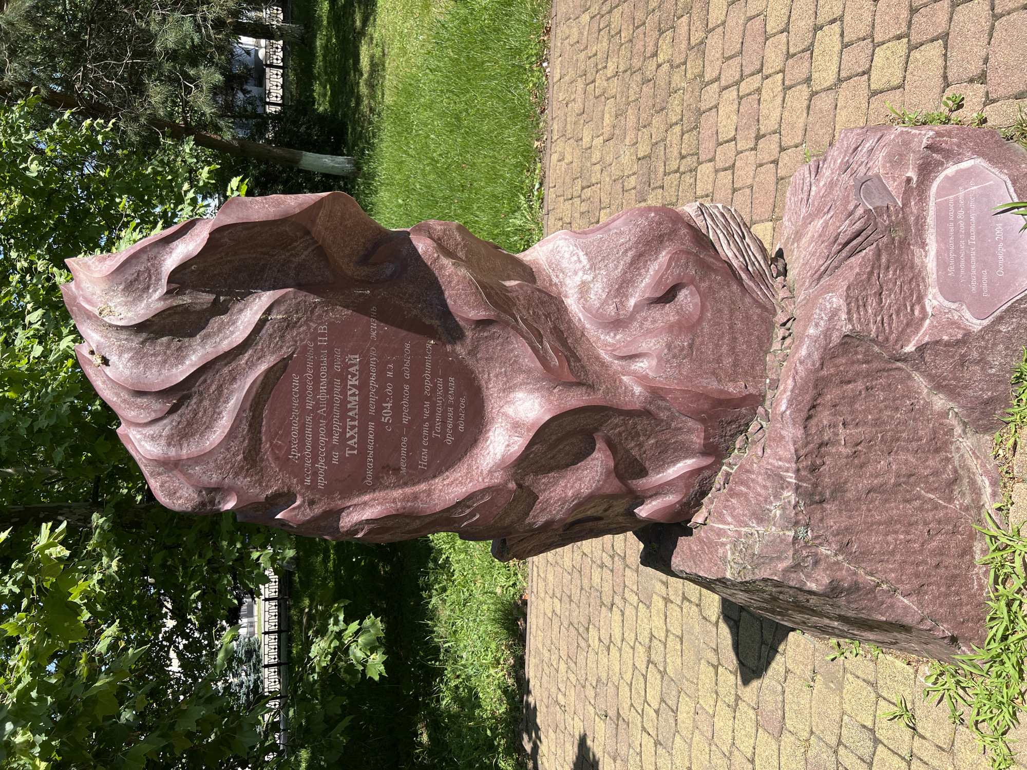 The image displays a large, abstract red stone monument with inscriptions, situated outdoors on a paved walkway in the city of Takhtamukai, Russia. The monument has a textured, sculpted surface resembling flowing rock or fabric, and it is supported by a larger, rougher stone base. The inscriptions on the monument are in Russian. To the left of the monument, a paved path made of rectangular yellow and reddish-brown bricks is visible. Behind the monument, a grassy area with lush green grass and trees with dense foliage can be seen. The trees are a mix of deciduous and coniferous. In the background, beyond the trees and grass, parts of a building and a decorative metal fence are faintly discernible. The lighting suggests a sunny day with clear skies, casting shadows from the monument onto the pavement. There are no people visible in the image. The overall scene depicts a monument in a park-like or public space within the city.