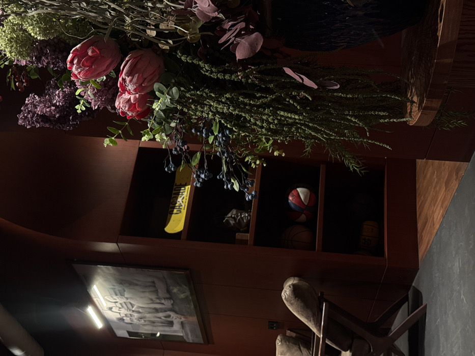 A floral arrangement with pink and purple flowers and greenery is atop a dark wooden shelf. On the shelf are a Cascade paddle, several balls, and small decorative objects. Below the shelf is a framed picture and a wooden chair. The chair has a carved back and a woven seat. The wall appears to be dark gray.