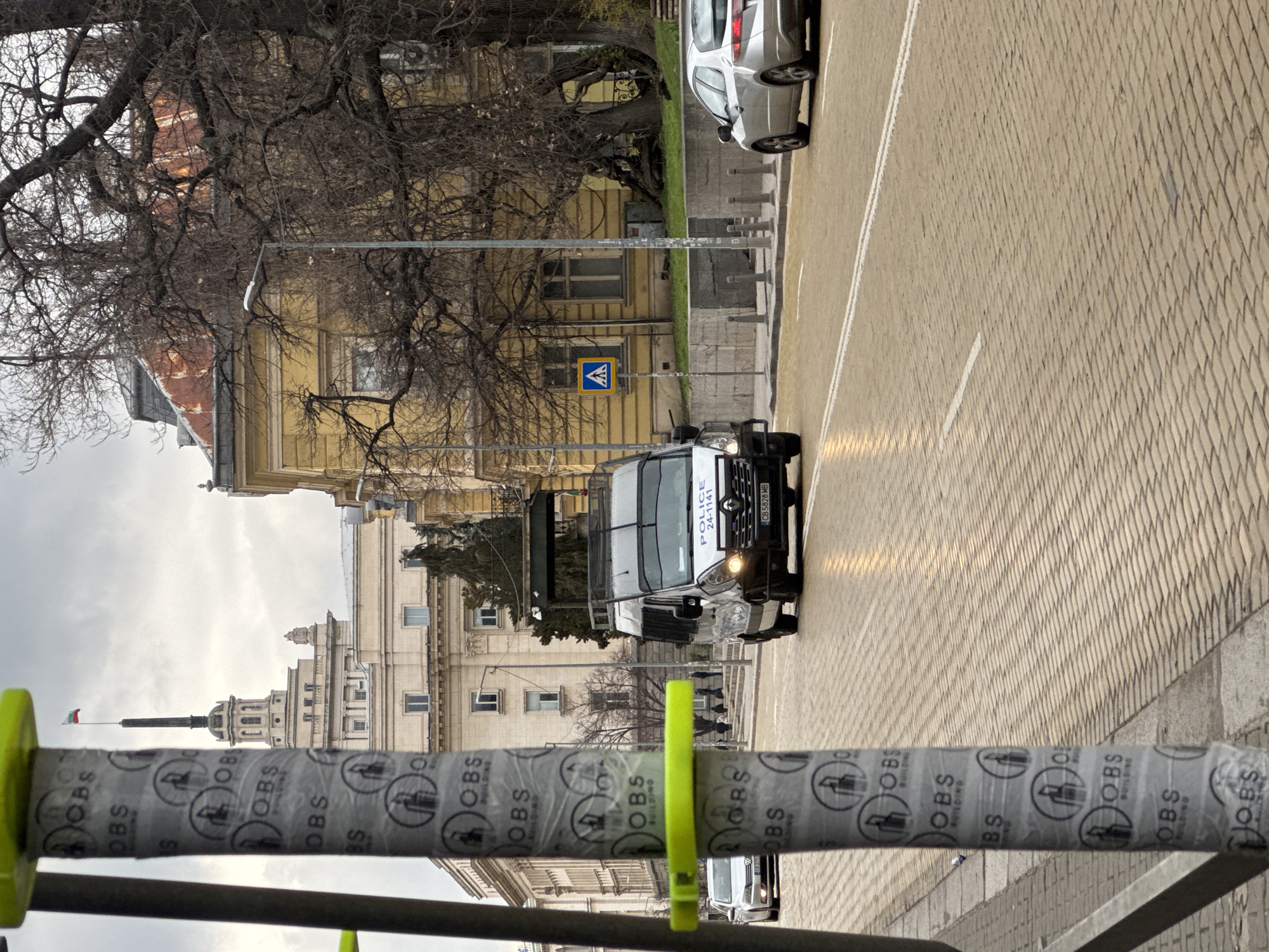 The image captures a street scene in Sofia, Bulgaria, from an eye-level perspective. In the foreground, a metallic railing is wrapped in a branded paper with "OBS BUILDING" printed repeatedly. Behind the railing, a police van is traveling on a brick-paved road. Street markings and other cars are visible. In the background, an assortment of old buildings are visible. A flag is visible atop the skyline in the distance. The sky is overcast.