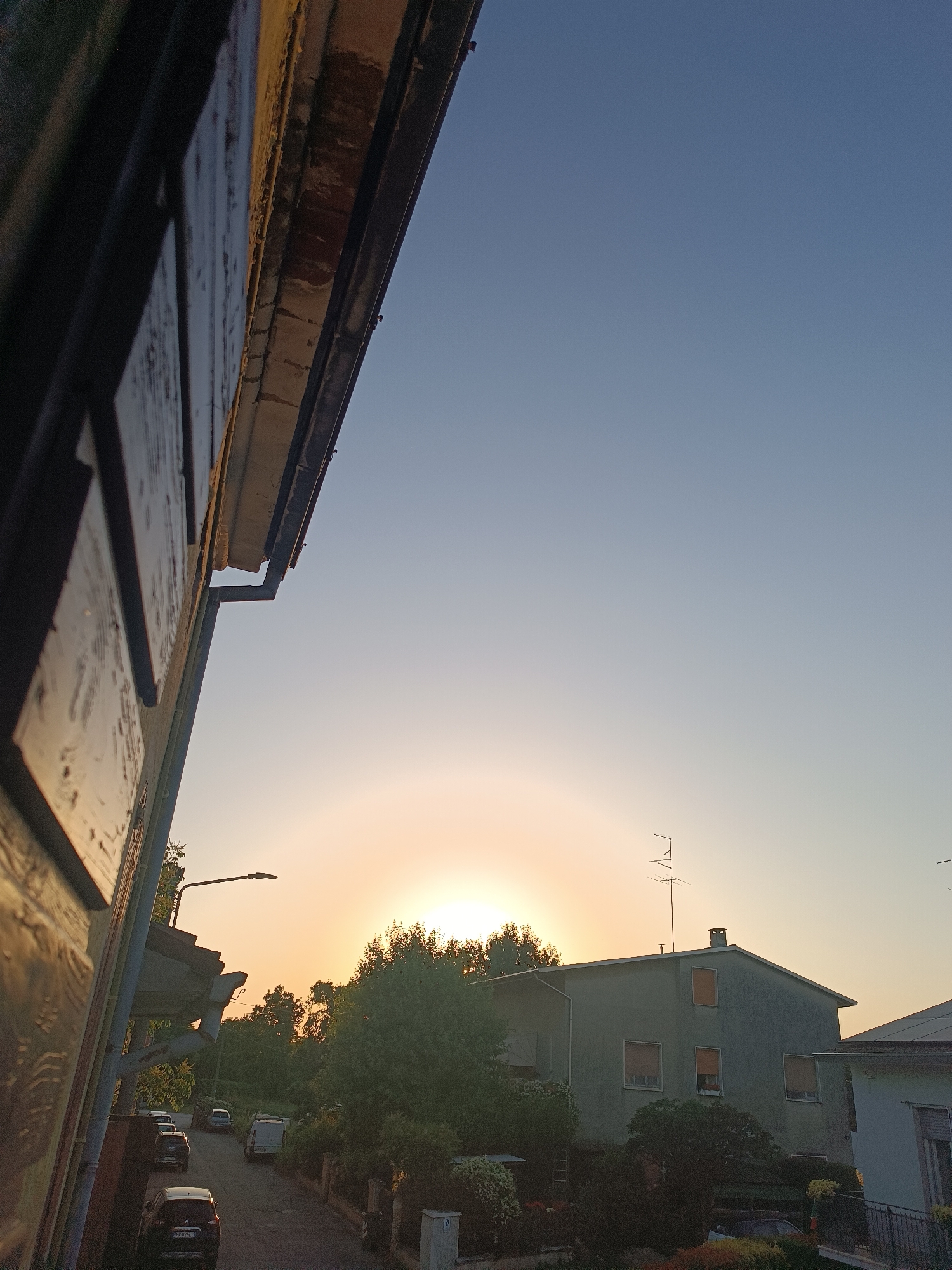 The image shows a residential street in Viadana, Italy, at dawn or dusk. The sky is transitioning from dark blue to orange near the horizon. A faint halo effect is visible around the sun. A two-story building with an antenna on its roof is situated in the middle ground. A row of trees partially obscures the sun. In the foreground, a section of a building with visible wooden eaves is on the left. Cars are parked along the side of the road. A street light is also visible.