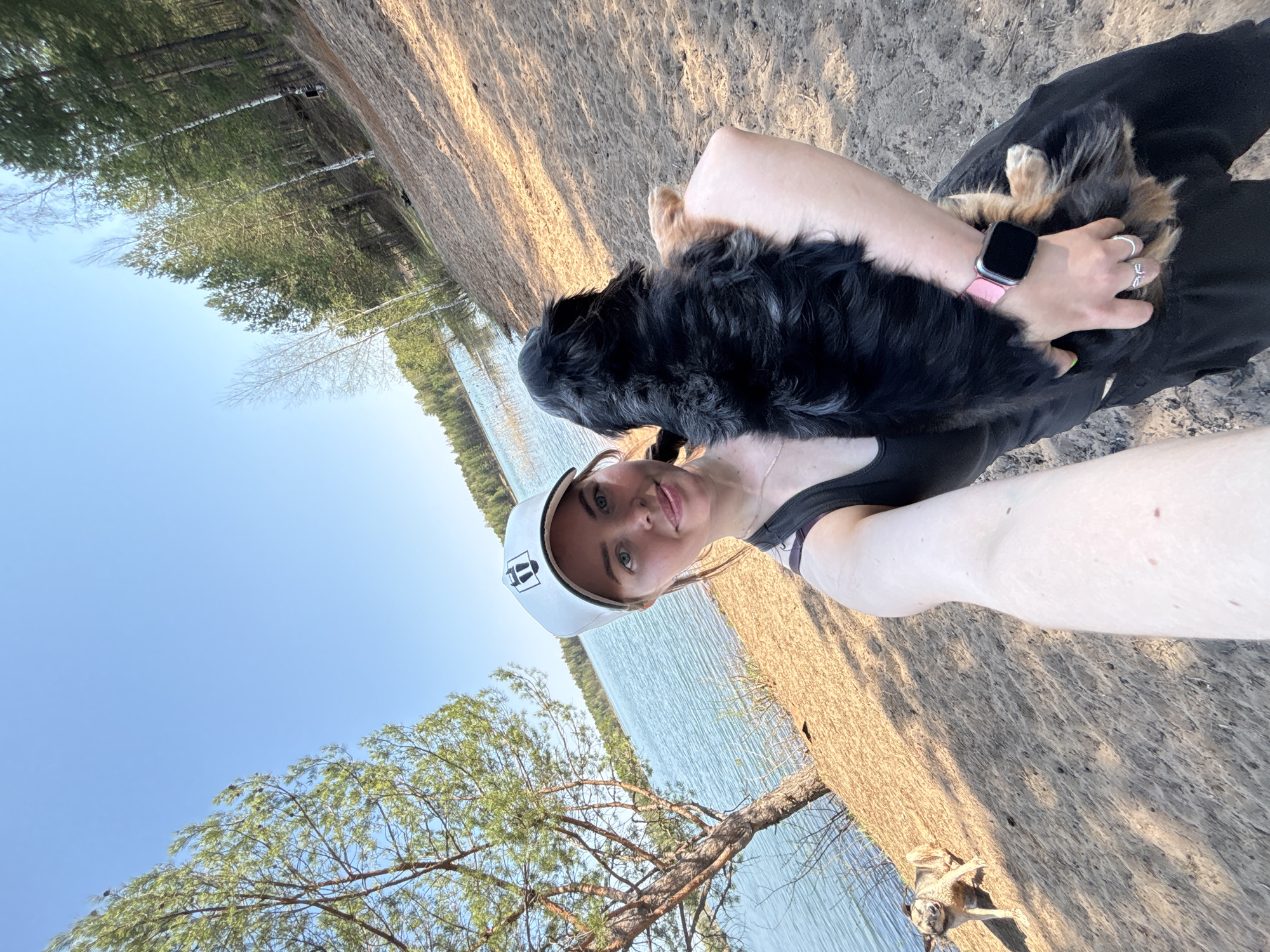 The image depicts a woman holding a small dog, likely a Bernese Mountain Dog puppy, outdoors near a body of water. The woman wears a white cap, a dark tank top, and a pink smartwatch. She is facing the camera and smiling. The dog is mostly black with some brown markings, held close to her chest. The background includes a shoreline with sandy ground, trees, and the surface of a lake or wide river. The sky is blue. The location appears to be a natural setting, but there is no specific evidence to confirm it is in Selychka, Russia. There is no evidence of urban development in the immediate vicinity. Another dog is visible in the bottom right corner of the image.