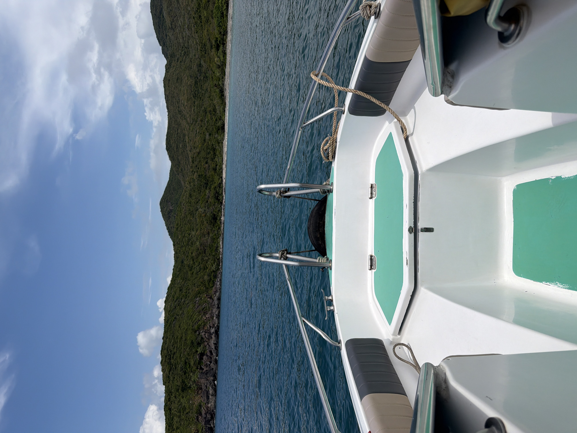 From the vantage point of a boat, a view encompasses the sea, a coastline of verdant mountains, and a partly cloudy sky. The boat's deck, painted white with green accents, is visible, featuring metal railings and a rope tied off to a cleat. The sea is deep blue and slightly choppy. The mountains are densely covered with vegetation, appearing as a solid green mass. The sky is a mix of clear blue and scattered white clouds. The location is Nha Trang Ward, Vietnam, inferred from geographical context.