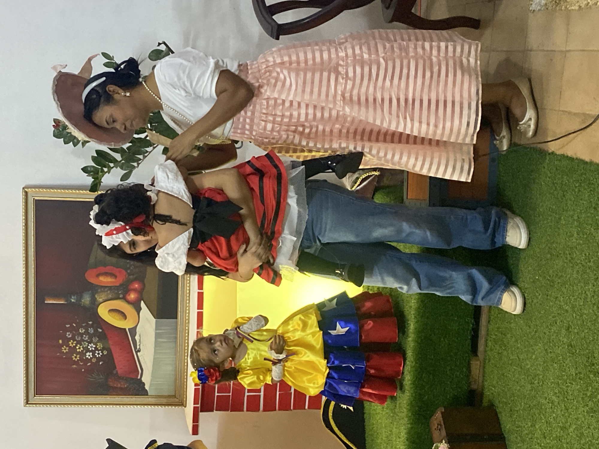 The scene depicts three females gathered indoors, possibly at a cultural event in Tinaquillo, Venezuela. The environment features a white wall with a large, framed still-life painting centrally mounted. The painting, gold-framed, portrays a dark bottle, a vase of white and yellow flowers, a slice of watermelon, a halved yellow melon, and red apples arranged on a white cloth against a deep red background. Below the painting is a decorative structure resembling a red and white brick fireplace mantel, with a yellow light source emanating from its opening.
To the left, a young girl with dark hair, styled with a Venezuelan flag-colored hair accessory, stands on green artificial turf. She wears a voluminous yellow dress with lower tiers in blue and red, adorned with white stars, mimicking the Venezuelan flag. Her forearms are covered with white lace armlets, and she holds a small, multi-colored ribbon. She is looking upwards and to the right.
In the center, an adult female with dark hair wears blue jeans and a white top. She is holding a smaller child, whose back is largely to the viewer. This child is dressed in a red outfit with a white top and a black bow at the back of the waist, with dark tights and shoes.
To the right, an adult female with dark hair pulled back into an updo, adorned with a light pink wide-brimmed hat, stands on a light-colored tiled floor. She wears a white short-sleeved top, a long, tiered light pink skirt with horizontal stripes, a pearl necklace, and silver ballet flats. She holds a small, metallic object near her chest with her left hand. Behind her, a leafy green plant is visible. A dark wooden chair is partially visible on the far right. A dark power cord extends across the tiled floor near her feet. A small dark wooden chest and a black pirate hat are positioned on the green turf in the lower-left foreground.