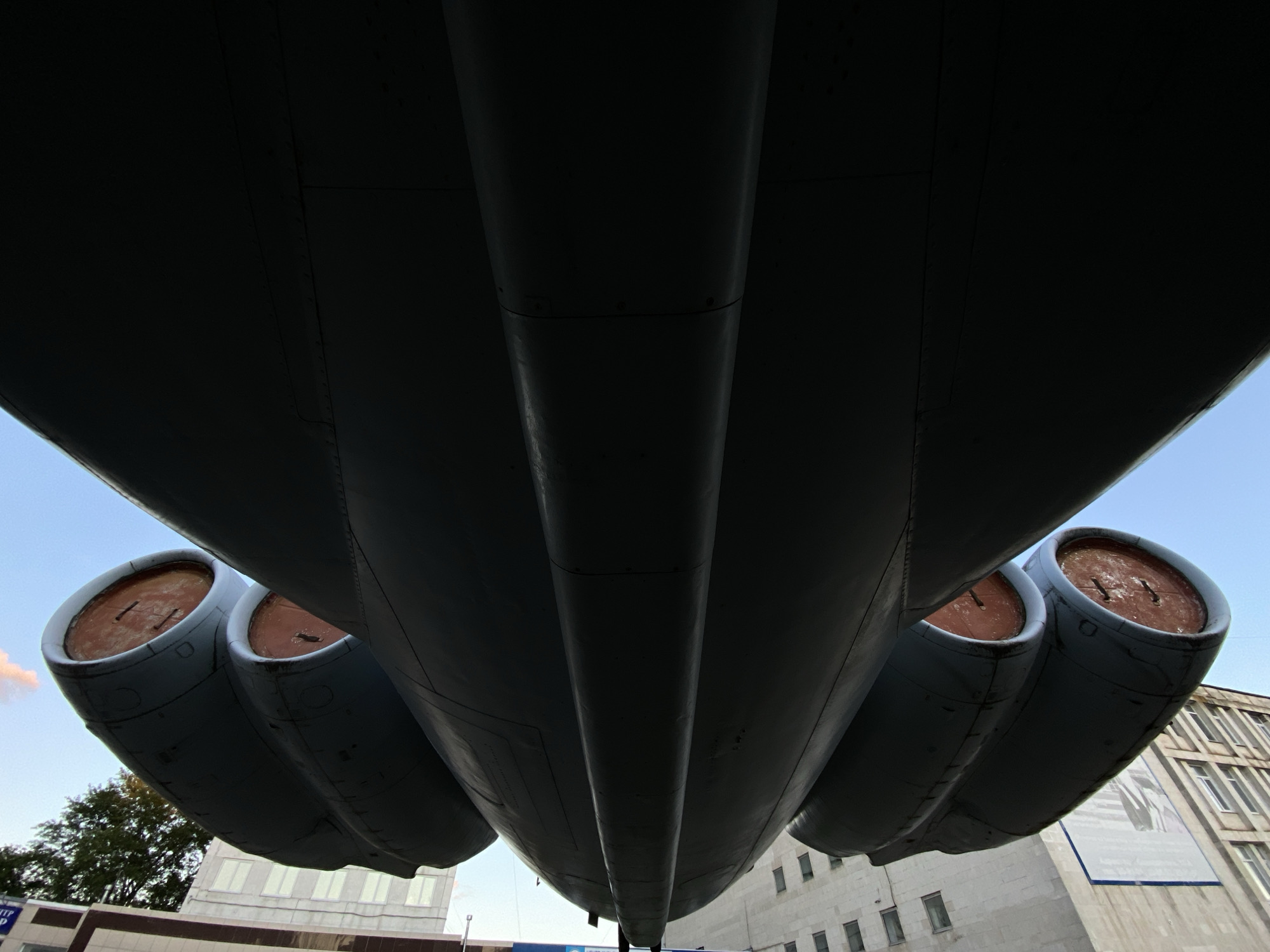 The image shows the underside of an aircraft's wing and engine nacelles, taken from a low angle. The wing is dark gray and dominates the upper portion of the frame, with visible panel lines and rivets. Two engine nacelles are visible in the foreground, each containing a circular inlet with a reddish-brown interior. The nacelles are cylindrical and appear to be made of metal.
In the background, a light-colored building with multiple windows is visible, suggesting an urban or industrial setting. Trees can be seen to the left of the building, and a clear blue sky is visible in the upper right corner.
This image was taken in Ulyanovsk, Russia, at the Central Museum of Civil Aviation, located at 104 Moskovskoye Shosse. The aircraft depicted is a Tupolev Tu-144, a supersonic transport aircraft.