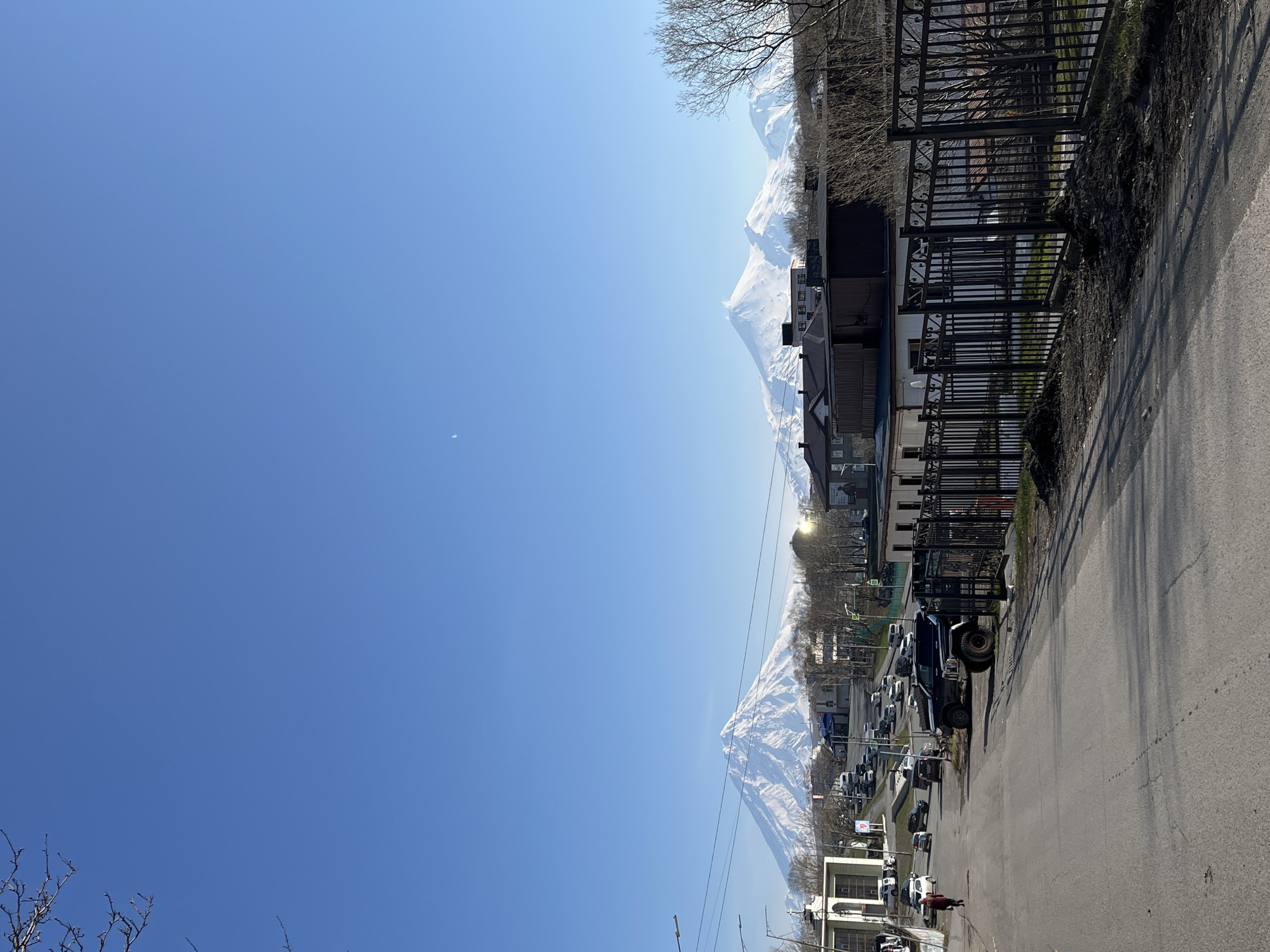 The image depicts a street scene in Petropavlovsk-Kamchatsky, Russia, under a clear blue sky. Snow-capped mountains are visible in the background. A road runs along the right side of the frame with parked cars lining the street. A black fence separates the street from buildings. Trees are also present, some bare of leaves. Pedestrians can be seen in the distance.
