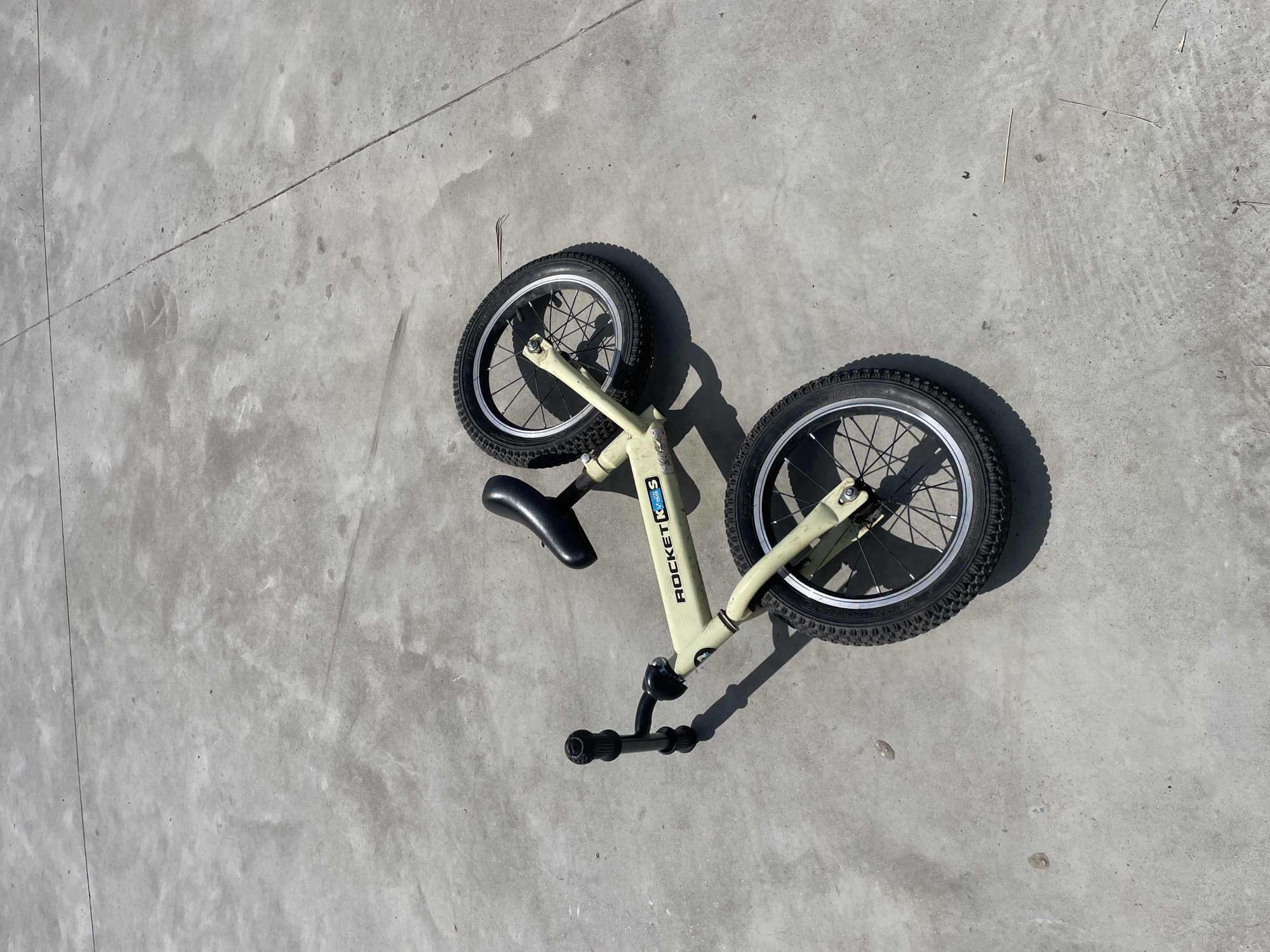 A children's balance bike, predominantly cream-colored with black handlebars, seat, and wheels, lies on its side on a cracked concrete surface. The bike features "ROCKET KS" branding in blue and white on the frame. The background consists of a light gray, textured concrete ground with some minor stains and debris. The scene appears to be outdoors, under bright sunlight, as evidenced by the shadows cast by the bicycle. The image was captured from a high angle, looking down at the bike. There are no people or animals present in the image. The location is Laishevo, Russia.