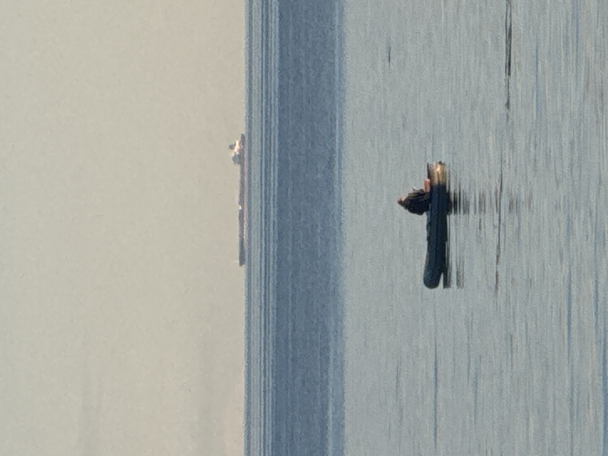 The image depicts a wide expanse of calm water, likely the Gulf of Finland, near Fort Krasnaya Gorka, Russia. The horizon line is clearly defined, separating the muted blue water from a pale, hazy sky. In the middle distance, a large, elongated cargo ship is visible, its silhouette a dark stripe against the lighter sky. Closer to the foreground, a small, dark inflatable boat is floating on the water. A single person, seen from behind, is seated in the boat, appearing to be fishing or simply observing the surroundings. The water is mostly smooth, with subtle ripple patterns that suggest a gentle breeze. The overall atmosphere is one of quiet solitude and natural beauty.