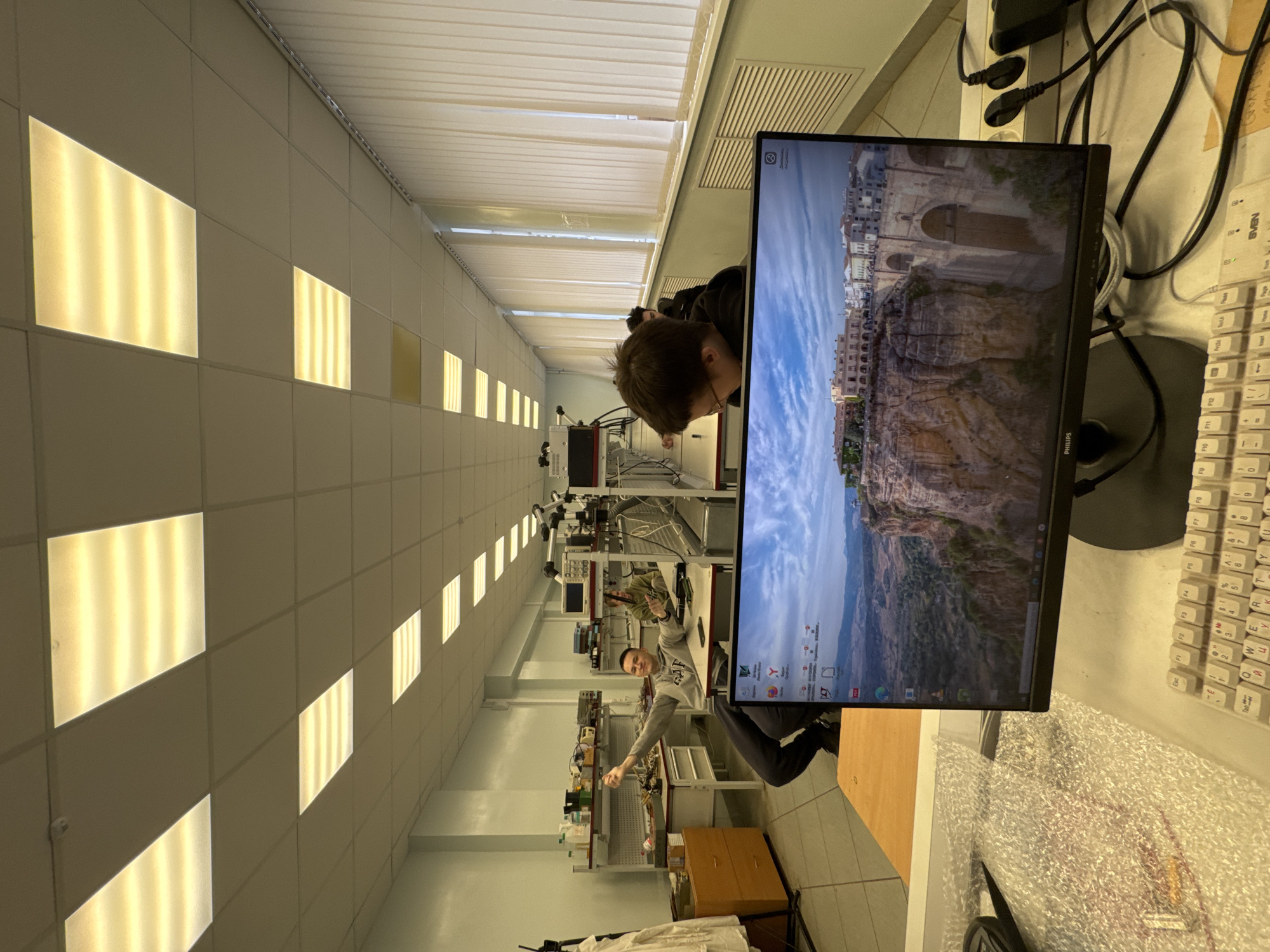 The image captures an indoor scene, likely a laboratory or workshop, with rows of fluorescent lights overhead and vertical blinds on the right side of the room. In the foreground, a computer monitor displays a landscape wallpaper of a city perched on a cliff. A white keyboard is visible in front of the monitor.
Further back, several individuals are present. One man, wearing a grey hoodie, is seated and appears to be gesturing with his right hand. He has short dark hair and is looking towards the camera. Another person, with darker hair and wearing glasses, is seated at a desk and looking down at the monitor. In the background, more desks and equipment are visible, suggesting a workspace. The overall environment appears to be a well-lit, functional space. There are no visible elements in the image that specifically indicate the city of Perm, Russia.