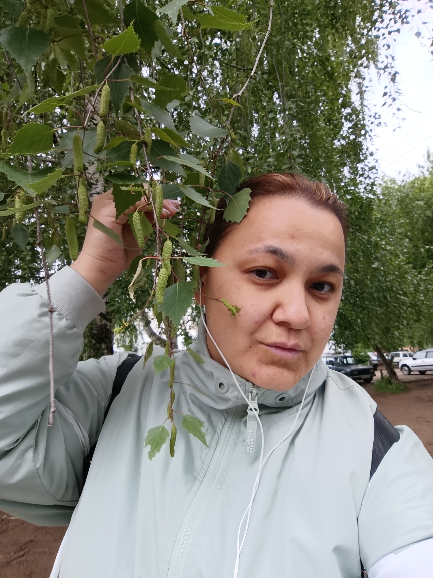 A woman stands outdoors near a birch tree in what appears to be a park or green space. She is wearing a light green jacket with dark gray accents, and white earphones are visible. She is holding a branch of the birch tree with leaves and catkins near her head. In the background, there are trees and parked cars. The location is likely in the city of Chaikovsky, Russia.
