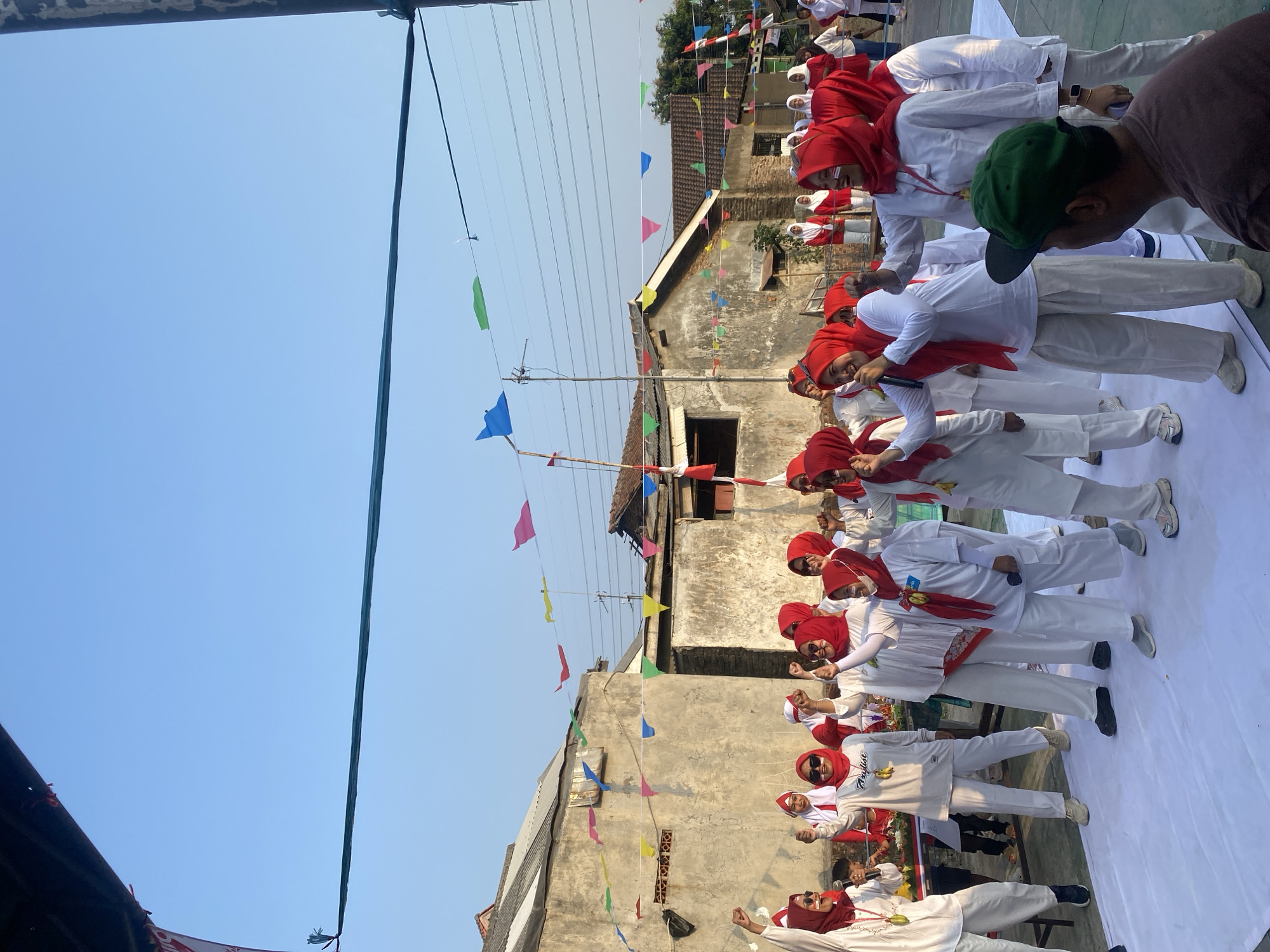 In a residential area of Cilegon, Indonesia, a group of women, predominantly dressed in white and red attire, participates in an outdoor event. The women are wearing red hijabs and some have white paint on their faces, suggesting a festive or patriotic occasion. They are standing on a white tarp spread on the ground, and many are raising their fists in the air, exhibiting a sense of excitement or solidarity. Some are holding microphones, indicating a performance or speech. In the background, simple, weathered buildings with tiled roofs are visible, adorned with colorful triangular flags strung across wires. The sky is clear and blue, suggesting it is daytime. A few spectators, including a man wearing a green baseball cap in the foreground, are observing the event. The overall atmosphere appears to be one of communal gathering and celebration.