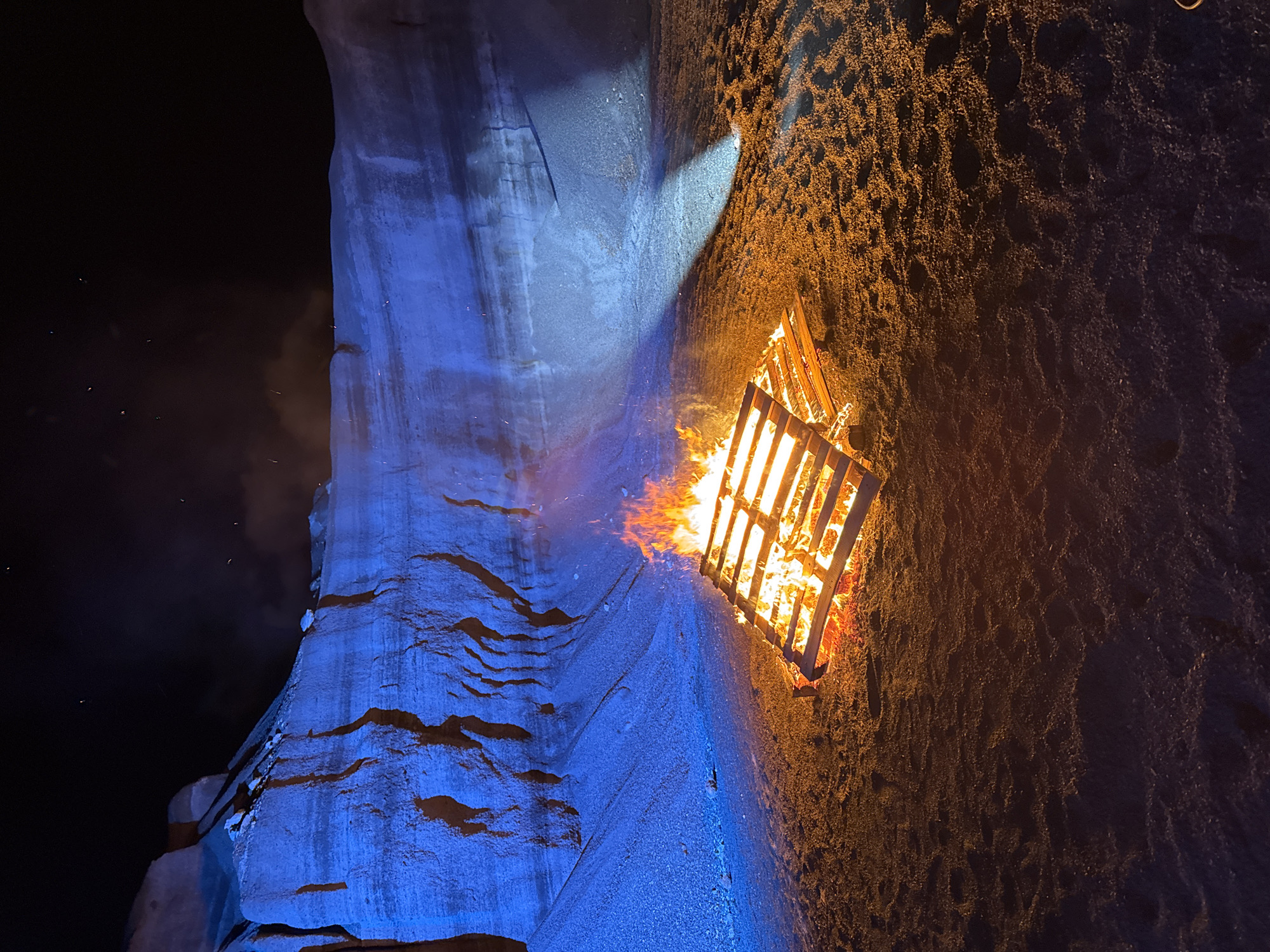 The image depicts a bonfire constructed from a wooden pallet, burning intensely on a sandy surface next to a rocky, cliff-like formation illuminated by blue light. The location is Teguise, Spain. The background shows a dark night sky.
