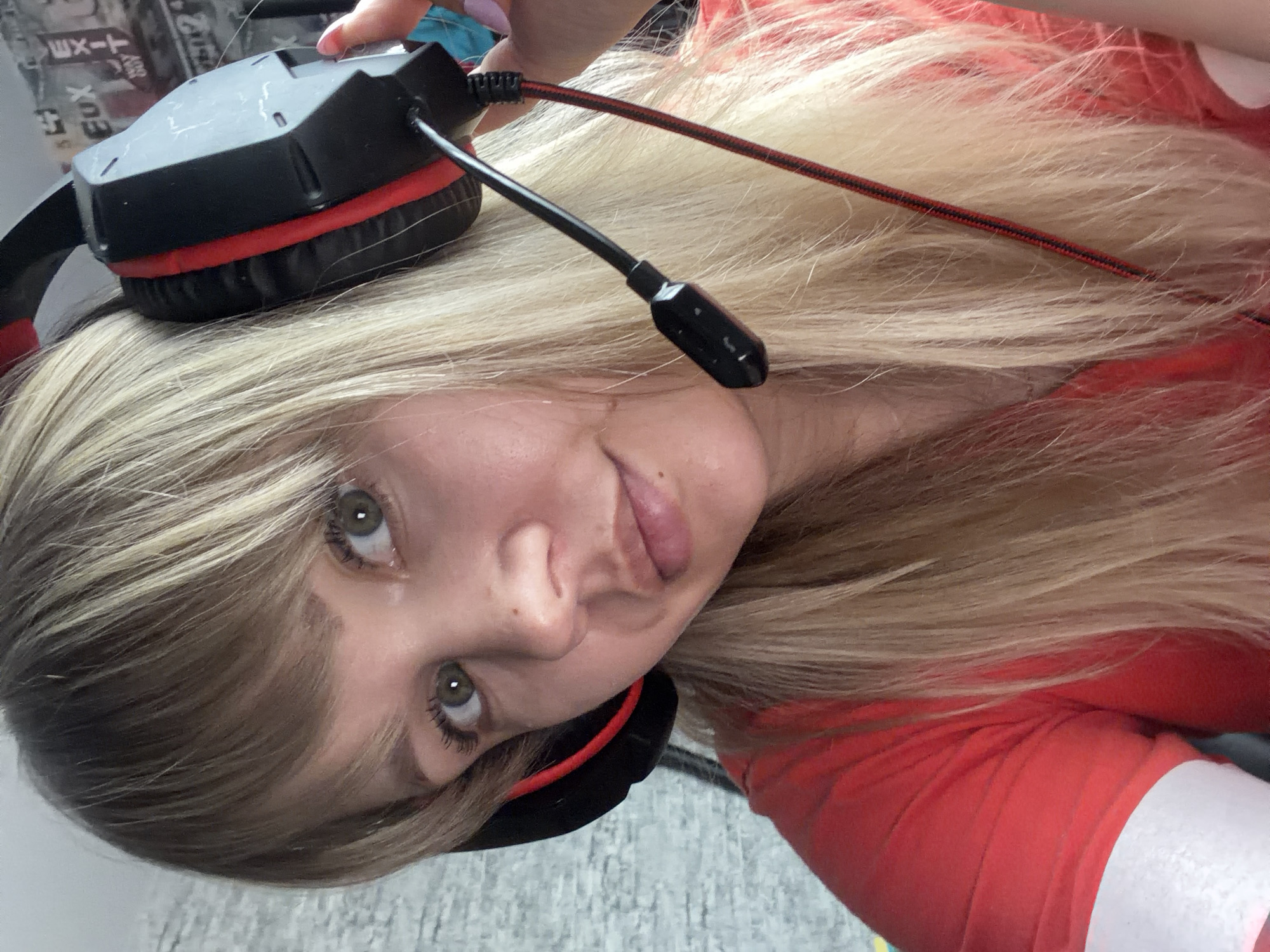 A close-up, tilted selfie shows a young woman with long, light blonde hair and light-colored eyes (likely green or hazel). She is wearing a black and red gaming headset, with one ear cup positioned over her right ear and the other resting on the top of her head. A microphone boom extends from the headset near her cheek. She wears a red long-sleeved top, with a white cuff visible on her right arm. Her expression is a subtle, playful half-smile, with her tongue slightly protruding from her mouth as she looks upward and to the viewer's left. The image is taken from a high angle, looking down at her.
The background is indoors and somewhat blurry. To the upper left, there are posters or papers on a wall, with partially visible black text that appears to include "EUX". Below her head, a light-colored, textured surface resembling a floor is visible. A red and black braided cable, likely associated with the headset, runs across her hair. The lighting suggests an indoor setting, possibly during the daytime.