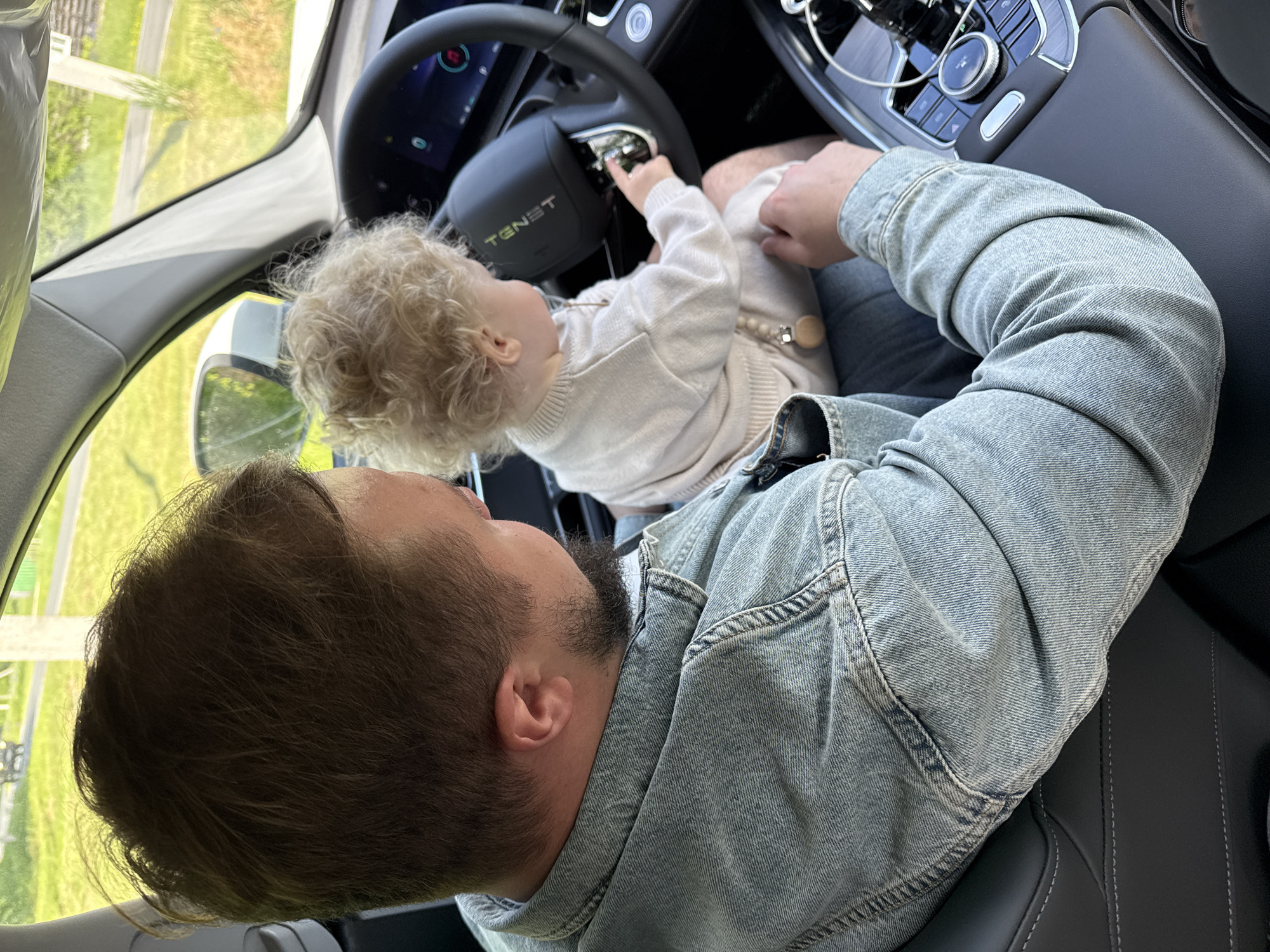 The image presents a top-down view inside the driver's side of a modern car, likely in Красный Маяк, Russia, during daytime. A man with dark hair and a beard, wearing a light blue denim jacket and blue jeans, is seated in the driver's seat. A young child, with curly blonde hair and wearing a light-colored long-sleeved top and pants, is sitting on the man's lap.
The child is actively engaged, reaching out with their right hand to touch the steering wheel or controls on the dashboard. The steering wheel features the prominent text "TENST." The car's interior is clean and features black leather seats with white stitching. The dashboard includes a screen, a central console with a rotary dial, and various buttons, suggesting a modern vehicle.
Through the car windows, a bright, sunny outdoor environment with green grass and some distant structures is visible, indicating the car is stationary or parked in an open area. A clear plastic film is present along the edge of the driver's side door frame, possibly a protective covering for a new vehicle. The scene depicts a moment of affectionate interaction and curiosity as the child explores the car's interior with the adult.