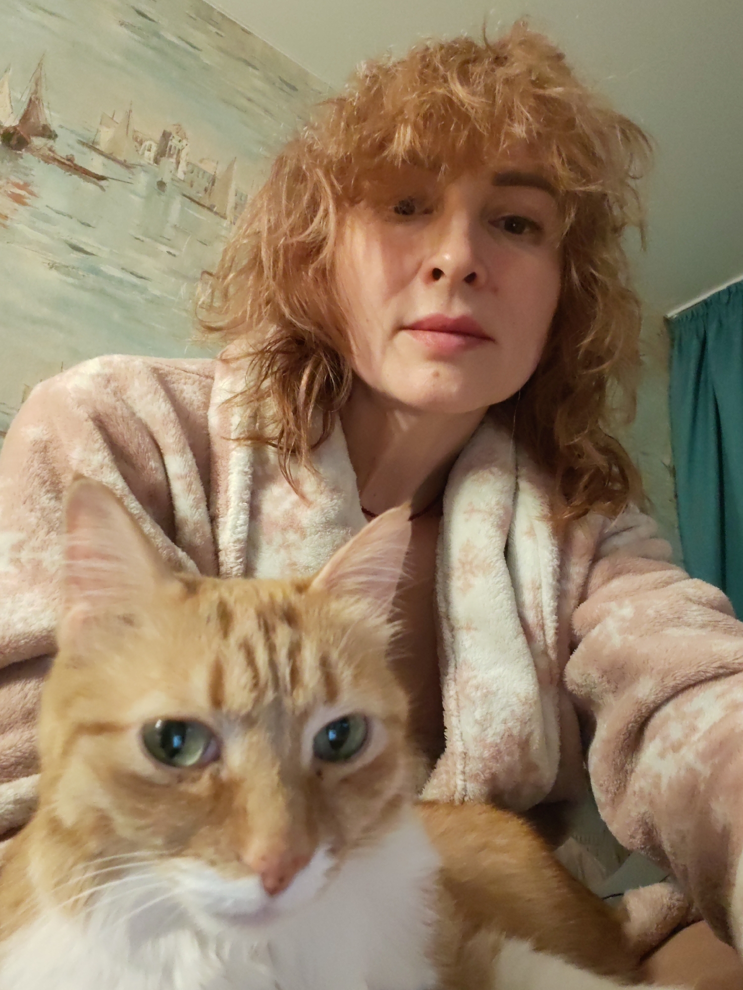 A woman and an orange tabby cat are featured in a close-up, selfie-style indoor photograph taken in Chaikovsky, Russia. The woman, with light, curly hair and bangs, is seen from the chest up, wearing a light pink or peach-colored fluffy bathrobe with a subtle pattern. She looks directly at the camera with a neutral expression. Positioned prominently in the foreground is an orange tabby cat with striking green eyes and white markings on its face and chest, also gazing towards the camera. The background reveals a wall adorned with a painting or wallpaper depicting a serene coastal scene with boats and buildings, possibly a European cityscape. A teal or dark green curtain is visible on the right side. The overall lighting suggests an indoor setting, likely a home environment. No text is visible.