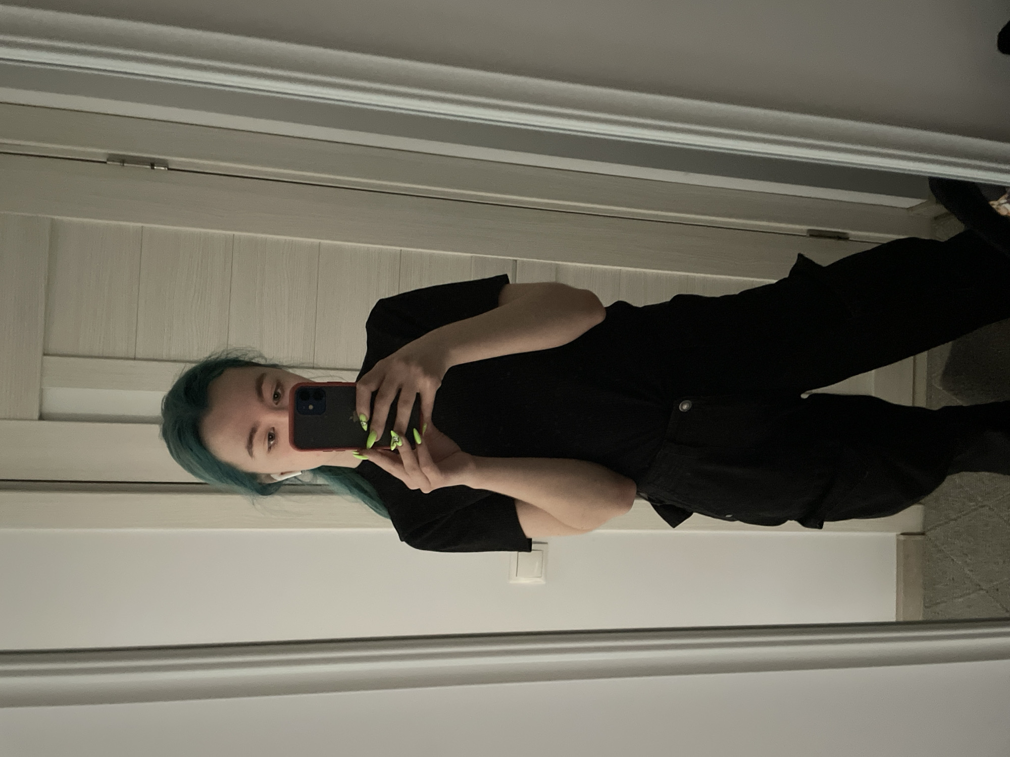 The media file shows a young person with teal-colored hair taking a mirror selfie. They are wearing a black t-shirt and black pants. The person is holding a smartphone in their right hand, capturing the reflection. Their fingernails are painted with bright green polish, some adorned with small designs. They are also wearing white earbuds. The background appears to be indoors, possibly in a room with white paneled walls and a white door or closet. The lighting suggests it might be daytime, though the exact time is not clear. The overall mood is casual and self-aware. The image provides a glimpse into a personal moment, focusing on the individual's appearance and the act of capturing their own image. No visible text is present in the image. The location context is Tallinn, Estonia.