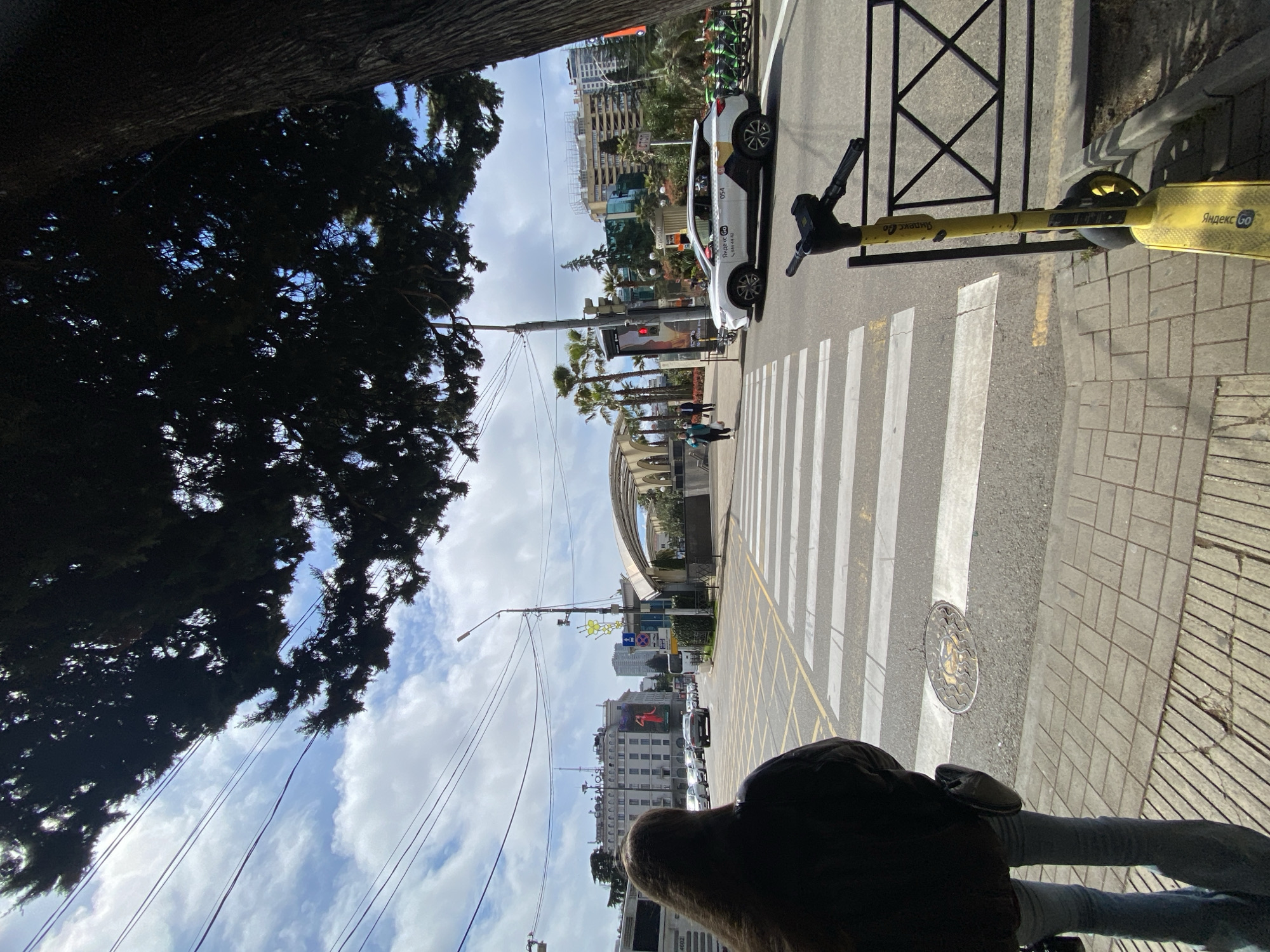 A person stands on the sidewalk adjacent to a crosswalk in Sochi, Russia. A yellow electric scooter leans against a black metal fence at the edge of the sidewalk. Across the street, a white car is parked near a line of parked bicycles. Palm trees and other vegetation line the street. Further in the background are buildings of varying heights and architectural styles. Overhead wires are strung across the sky, which is partly cloudy. The scene is partially obscured by a tree.