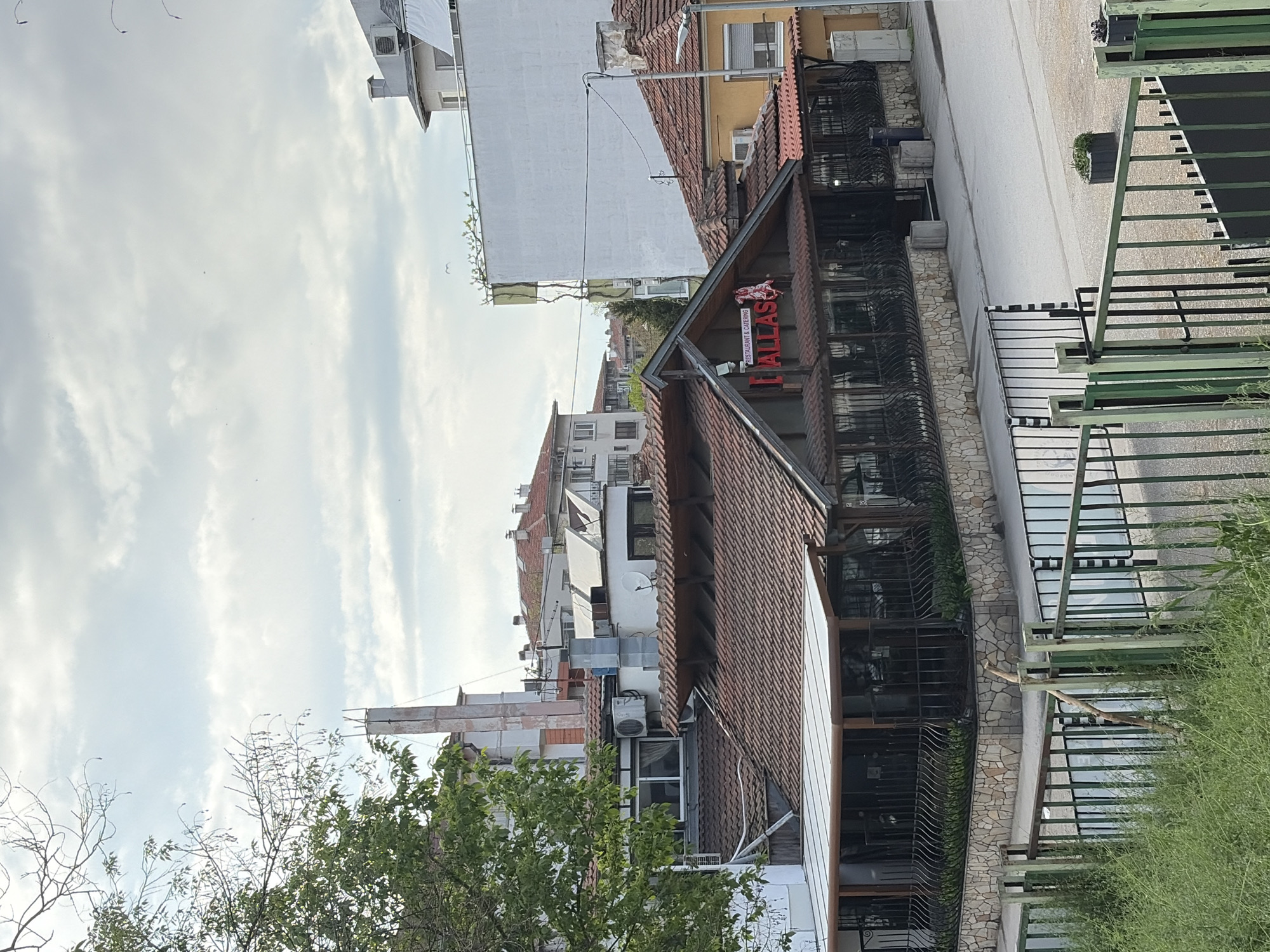 The image captures a daytime street scene in an urban environment, likely Plovdiv, Bulgaria. The sky is bright with scattered clouds, suggesting fair weather. No people are visible.
A prominent building on the right features a red illuminated sign that reads "DALLAS" in large capital letters, with "RESTAURANT & CATERING" written below it in smaller white text. A stylized animal emblem, possibly a bull or ram, is placed above "DALLAS." This building has a distinctive terracotta-tiled roof and large windows protected by ornate black metal grates. Its lower facade is clad with stone.
In front of the "DALLAS" building, a paved street runs horizontally across the mid-ground. The street appears empty, with a single potted plant near its center. To the right of the street, a green slatted fence or railing is partially obscured by green foliage and bushes in the foreground.
Beyond the main restaurant building, and extending into the background, are several other buildings with varied architectural styles, including multi-story structures with white walls and others with tiled roofs. Air conditioning units and a satellite dish are visible on some of these buildings, along with utility poles. Green trees and foliage are present in the foreground and various parts of the scene, adding natural elements to the urban landscape. The overall impression is that of a quiet street on a bright day.
