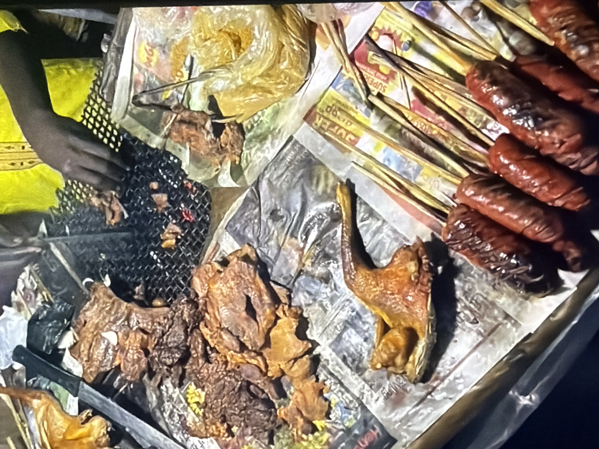 A close-up, eye-level shot shows a street food vendor in an outdoor setting, likely at night given the lighting. The foreground features several skewers of grilled sausages, which are dark red and appear slightly charred. To the left of the sausages, a whole roasted chicken or fowl is positioned, with its wings and legs splayed out. Behind the fowl, a large pile of grilled meat, possibly beef or lamb, is visible, interspersed with what looks like chunks of fat. The meat is arranged on what appears to be newspaper lining a food stall. In the background, a person wearing a bright yellow garment with gold trim is tending to a grill, their dark hands visible as they handle the food. A metal tong rests on a crumpled plastic bag filled with a yellow spice, possibly turmeric. The overall scene depicts a vibrant street food preparation area.