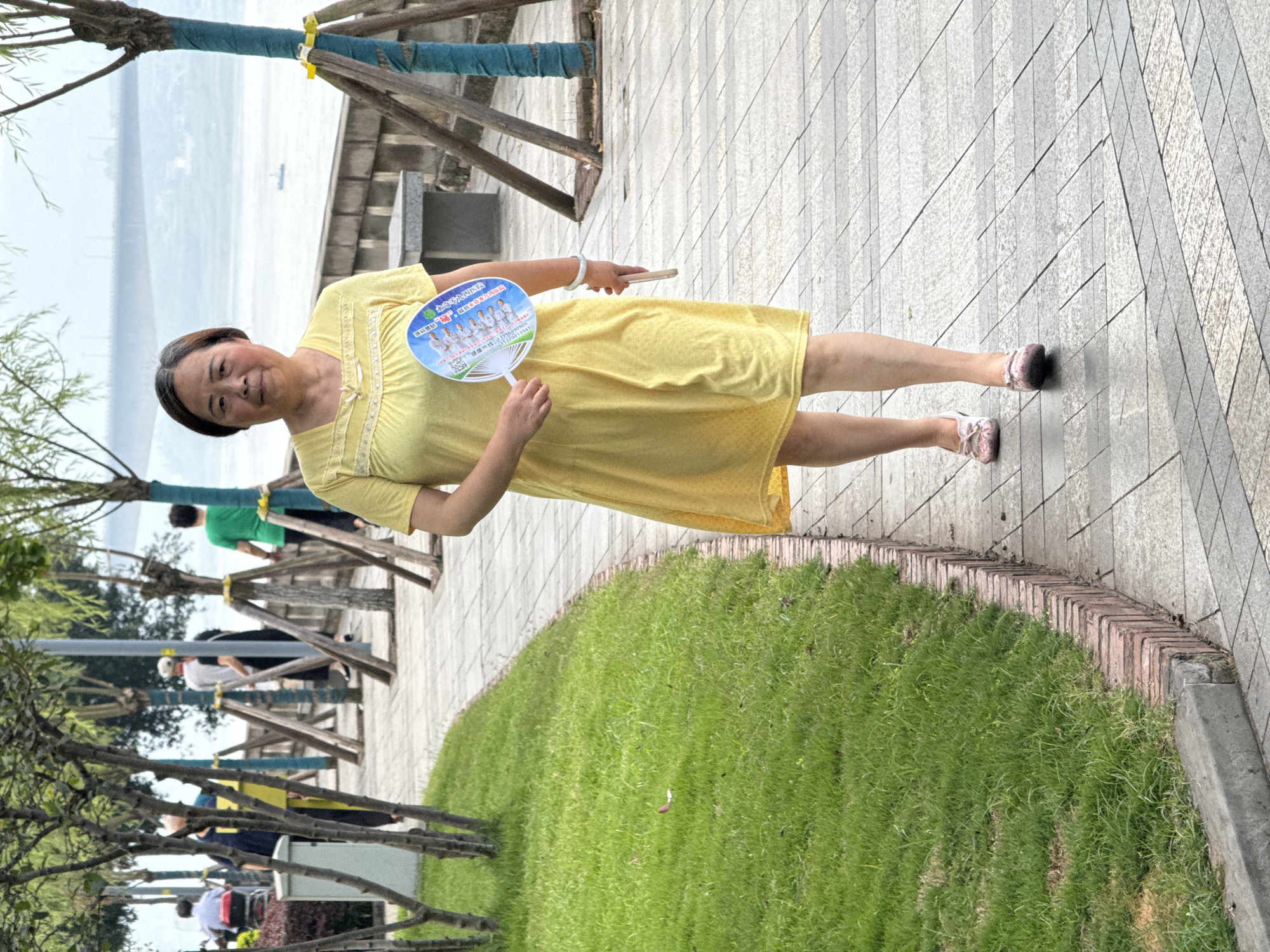 This image captures an outdoor scene in the city of Jiangjin, China, along a paved riverside promenade or park path. The sky appears overcast or hazy.
A middle-aged East Asian woman, with dark hair styled back, walks directly towards the viewer in the foreground. She wears a knee-length, short-sleeved yellow dress with a textured pattern and lace trim around the neckline. On her feet are light-colored, possibly pink, flat shoes featuring a decorative element on the toe. In her left hand, she holds a white and blue promotional fan displaying images of several individuals, Chinese text, and phone numbers. Her right hand holds a light-colored smartphone, and she wears a light-colored bracelet on her right wrist. Her expression is neutral as she looks forward.
In the background, several other individuals are visible further along the path. On the far left, a person is seated in a red-seated mobility device. Moving right, another person in a dark top stands near a yellow vertical object. Two people walk together, one in a white top facing away, and another in dark clothing walking beside them. Further right, a person in a bright green t-shirt walks away from the camera.
The walkway is paved with a pattern of light-colored, rectangular tiles in shades of grey and beige. To the left of the path, a strip of green grass is bordered by a low brick curb and a concrete curb. Numerous young trees with slender trunks line the path; many are supported by wooden stakes or frames, and some trunks are wrapped with blue fabric. Beyond the path and a low wall or embankment, a wide body of water, likely a river, stretches into the distance. A small boat is visible on the water. A large, hazy bridge spans the water in the far background. On the right side of the path, a concrete bench and a dark utility box are visible behind the main subject. A grey utility box is also present on the left side of the path near the grassy area.