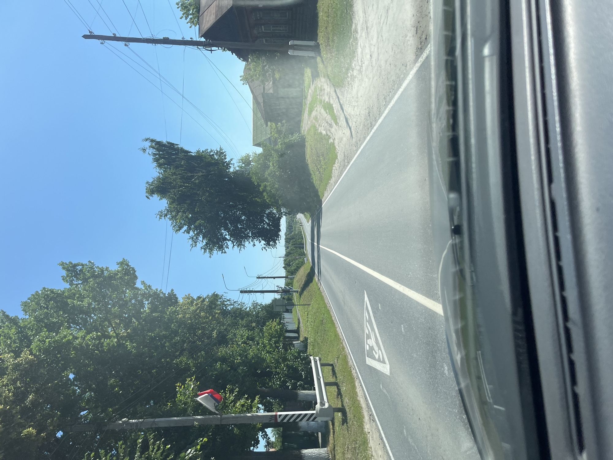 The image shows a view from inside a vehicle driving on a paved road in Yadrin, Russia. The road is flanked by trees and a grassy verge on both sides. On the left side, there is a utility pole with a small Russian flag attached, and a white metal guardrail. Further down the road on the left, a white fence with trees behind it is visible. On the right side of the road, there is a dirt path leading to several wooden buildings, one of which appears to be a house with ornate window frames. The sky is clear and blue. The road itself has white lane markings, and a triangular road sign depicting a child crossing is painted on the pavement in the foreground. The dashboard of the vehicle is visible at the bottom of the frame. The overall impression is a rural or semi-rural setting on a sunny day.