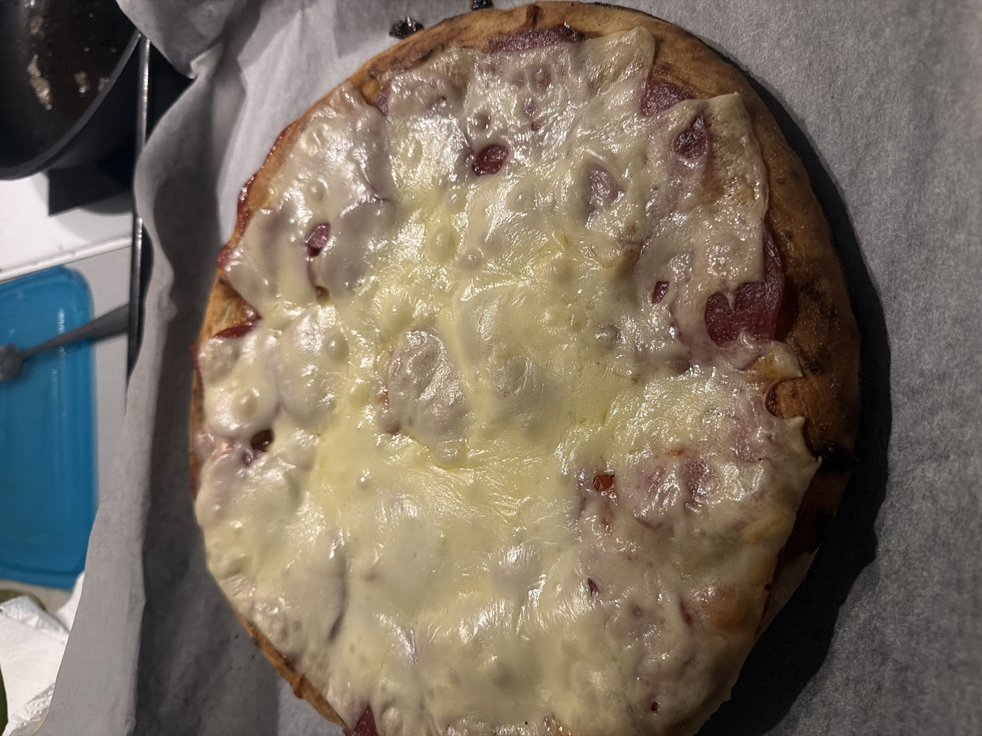 The image shows a close-up, overhead view of a partially baked pizza with melted cheese. The pizza has a golden-brown crust and appears to have red toppings visible beneath the cheese. It is placed on a baking sheet lined with parchment paper. The background is blurred but suggests a kitchen environment with some cooking utensils and a blue object, possibly a container. There are no people or animals visible in the image. The scene does not provide any specific details that would allow for a location to be identified in the city of Szczecin, Poland.