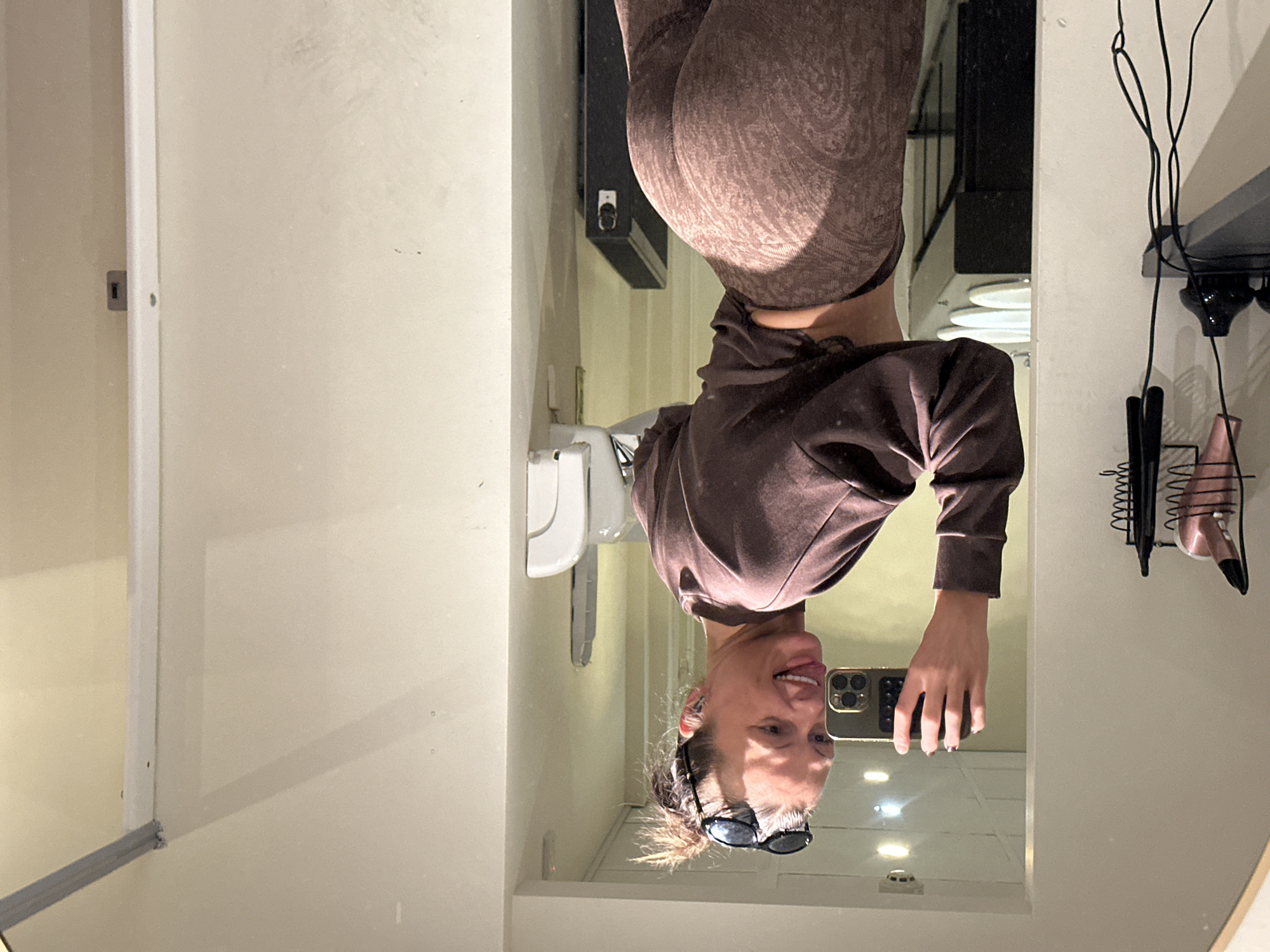 The image is a mirror selfie taken indoors, rotated 90 degrees clockwise, showing a person appearing upside down. The subject, an adult with brown hair tied back and black sunglasses pushed up on their head, is smiling broadly with their tongue slightly extended. They are holding a smartphone, which appears to be an iPhone, to capture the reflection.
The person is dressed in a dark brown long-sleeved top and dark brown leggings featuring a subtle, lighter-toned paisley-like pattern. The reflection in the mirror reveals a room with light-colored walls, possibly off-white or beige. In the background of the reflection, a white toilet or sink fixture is visible. To the right, a black shelf or cabinet is mounted on the wall, alongside a black metal wire organizer holding various items, including what looks like a pink hairdryer and other tools. The lighting suggests an indoor setting with artificial illumination. The image captures a casual and playful moment. No discernible text is visible.