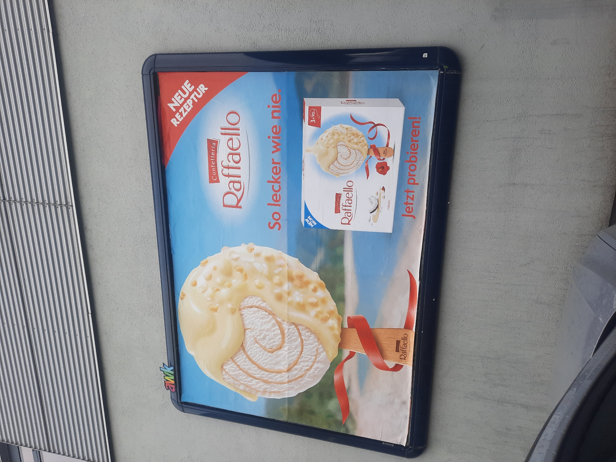 The image shows a close-up of an outdoor billboard advertisement for Raffaello ice cream. The billboard is mounted on a textured, light gray wall. Above the billboard, a section of a modern building with horizontal gray siding is visible. The billboard itself features a large image of a Raffaello ice cream cone with white chocolate and nuts, partially covered in a red ribbon. To the right of the ice cream cone is a box of Raffaello ice cream. The text on the billboard includes "Confetteria Raffaello," "So lecker wie nie." (So delicious like never before), "NEUE REZEPTUR" (NEW RECIPE), and "Jetzt probieren!" (Try it now!). The background of the advertisement depicts a blurred scene of a beach and ocean. A small, colorful "awk" logo is present in the upper left corner of the billboard. The bottom portion of the image shows the hood of a dark-colored car, partially obscuring the lower part of the billboard and the wall below it.