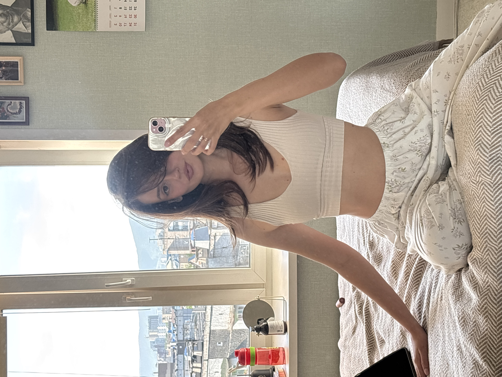 The image, rotated 90 degrees counter-clockwise, depicts a young woman taking a mirror selfie indoors during the daytime. She is seated or leaning on a bed or couch, wearing a light cream-colored ribbed crop top and loose, white trousers with a subtle floral pattern. Her dark hair frames her face, and she holds a light-colored smartphone with a clear case and multiple camera lenses, reflecting her partially visible face which shows a calm or slightly smiling expression. A ring is visible on her finger.
Behind the woman is a large window providing a panoramic view of a city with numerous buildings and distant mountains under a bright, clear sky, indicating clear weather. On the windowsill, several items are arranged, including a small round mirror on a stand, two bottles (one dark, one light), and two colorful reusable water bottles (red and green).
The interior wall above the window is a light green textured surface, decorated with framed pictures. These include a portrait of an older man, an image of a dog or lamb, and two smaller frames. A calendar displaying numbers like 1, 2, 3, 10, 11, 12, 18, 19, 23, 24, 25, 30, and 31 is also visible. The bed or couch is covered with a textured blanket featuring a chevron or herringbone pattern in light brown and white. A black tablet or laptop is partially visible on the blanket near her right hand, and a small lip gloss tube lies nearby.
The bright natural light suggests it is morning or early afternoon. The urban landscape with mountains is consistent with the provided location context of Krasnoyarsk, Russia.