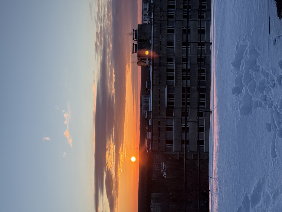 The image depicts a snow-covered urban landscape in Prypiat, Ukraine, at either sunrise or sunset. The foreground is occupied by a flat surface, likely a rooftop, blanketed in undisturbed snow with numerous visible footprints, suggesting recent human presence.
In the midground, a large, multi-story concrete residential building, typical of Soviet-era architecture, dominates the view. Its facade is weathered grey, featuring multiple windows, some dark and empty, others reflecting light. The building's roof features two prominent rectangular structures with rounded tops, possibly ventilation shafts, with small, bare saplings growing nearby. A distinct yellow-orange sun flare is visible on the side of the rightmost rooftop structure.
The background reveals a brilliant orange horizon where the sun, a bright circular orb, is partially visible, positioned slightly left of center. Above the horizon, the sky transitions to a pale blue, with horizontal cloud formations displaying shades of grey, pink, and orange. Below the horizon line, a dark band of silhouetted trees or distant urban structures extends across the landscape. Further into the distance, several more multi-story buildings, consistent with Prypiat's architectural style, are faintly visible against the sky. No individuals are observed in the scene. The overall environment suggests a cold, desolate setting.
