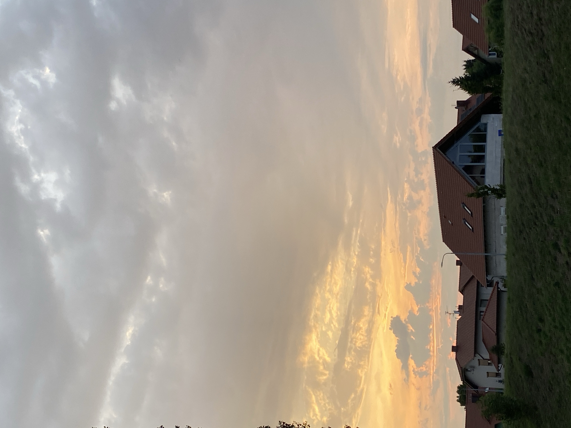 The image shows a residential area at dusk, likely in the city of Buk, Poland. The sky is filled with a mix of gray and orange clouds, indicating the setting sun. Several houses with red-tiled roofs are visible, nestled behind a grassy embankment. One house in the foreground has a distinctive triangular window. Trees and foliage are scattered throughout the scene. There are no people or animals present. The overall impression is one of a quiet, suburban evening.