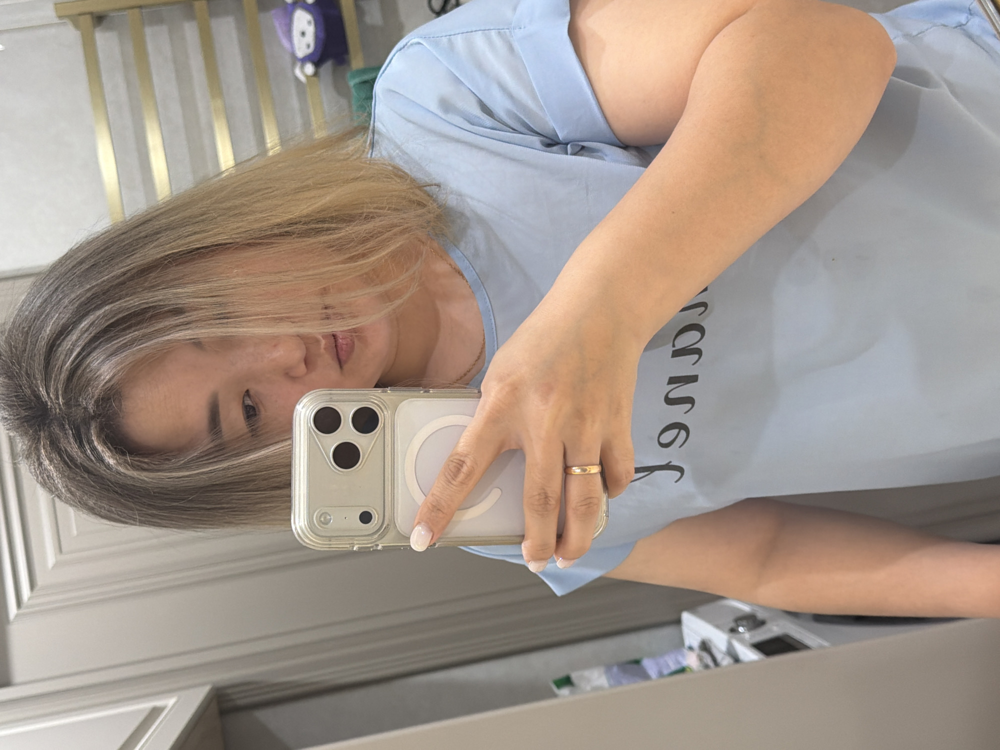 A woman with blonde hair is taking a mirror selfie in a bathroom. She is wearing a light blue t-shirt and a gold ring on her left ring finger. Her phone is visible in her hand. In the background, a gold towel rack with a purple stuffed toy is mounted on the wall. A washing machine is visible in the reflection of the mirror on the left side of the image. The overall environment appears to be a domestic bathroom in Astana, Kazakhstan.