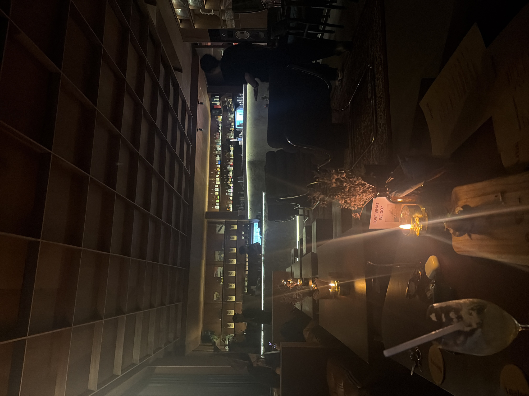 The image depicts the interior of a dimly lit bar with wooden shelving units lining one wall. The shelves are filled with bottles, suggesting a well-stocked bar. In the background, a bar counter is visible, with bottles displayed on shelves and a bartender possibly working behind it. There are several patrons seated at the counter and at tables. One table in the foreground has a lit candle, a small decorative plant, and a glass with a straw. A sign on the wall above the table reads "LOVE WHAT WE DO?". The overall ambiance is intimate and sophisticated, suggesting an evening setting. The lighting is warm and inviting, with spotlights illuminating the bar area and accent lighting along the shelves. The scene appears to be in a bar or lounge in North Kuta, Indonesia.