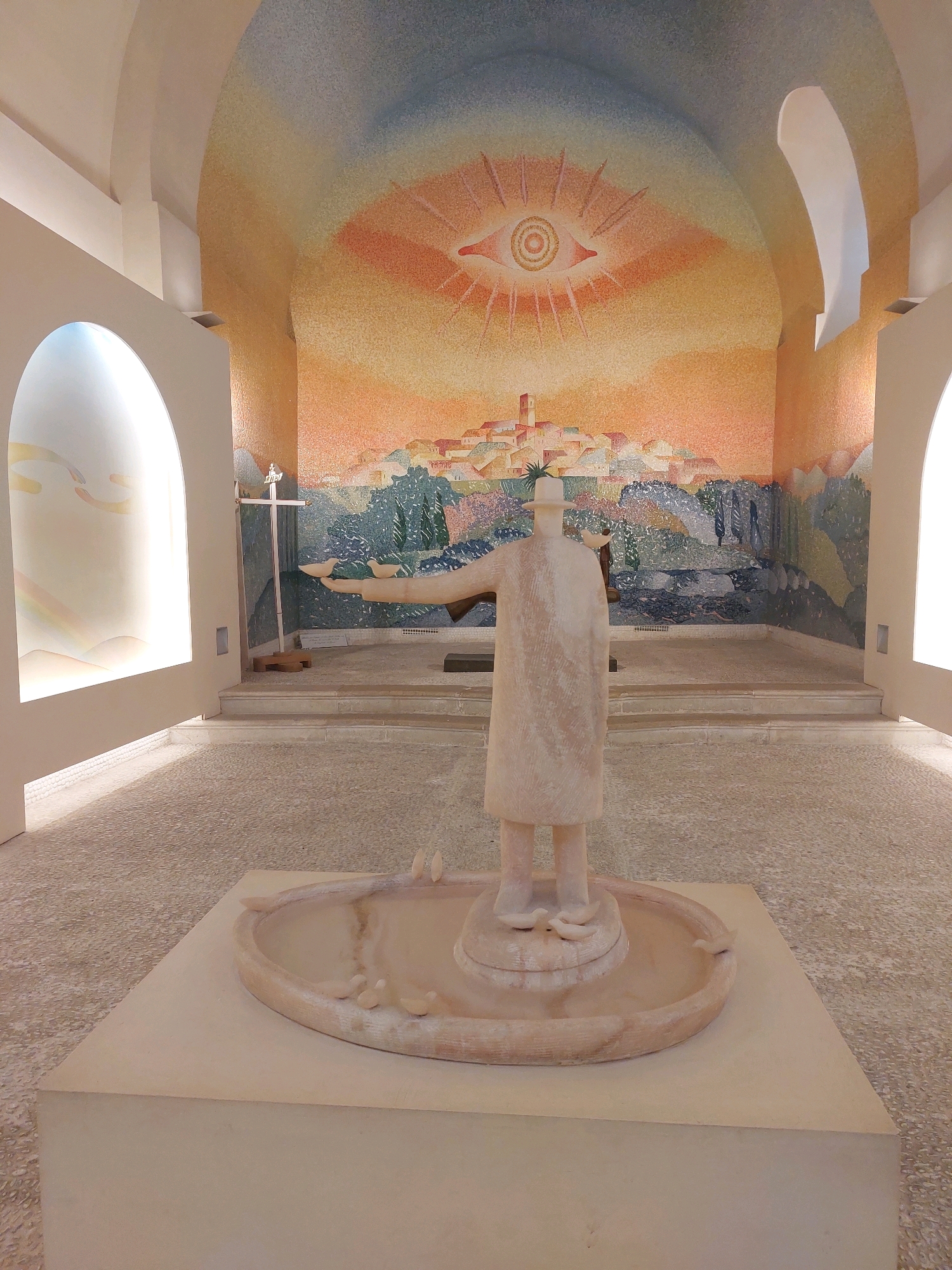The image depicts the interior of the Fondation Maeght in Saint-Paul-de-Vence, France. The central focus is a pale stone sculpture of a figure wearing a hat and a long coat, with arms outstretched. The sculpture stands on a circular stone base that appears to be a fountain, with small bird figures placed around its rim and on the figure's outstretched hand. Behind the sculpture, a large mosaic mural adorns the curved wall, depicting a stylized landscape with a village and a prominent eye symbol radiating light. To the left, an arched alcove with soft lighting creates a serene ambiance. The floor is paved with small, light-colored stones. A cross with two dove figures is visible to the left of the sculpture. The overall scene suggests a tranquil and artistic environment.