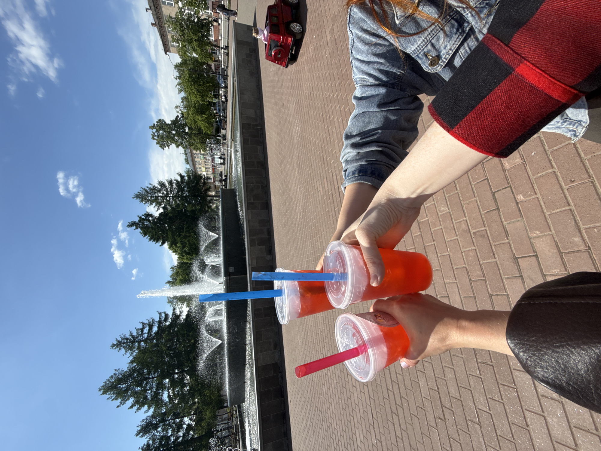 Two people are holding up three drinks in plastic cups with straws. One drink has a red straw and two have blue straws. The drinks are filled with an orange-colored liquid. One person is wearing a blue denim jacket with a red and black flannel sleeve. The other person is wearing a black jacket. In the background, there is a fountain, trees, buildings, a red children's car, and a blue sky with some clouds. The location is likely Chaikovsky, Russia, based on visual cues.