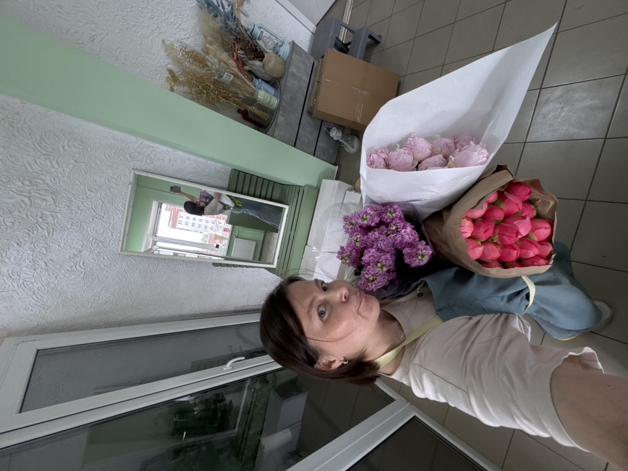 A selfie taken in what appears to be a florist's shop or a room with floral arrangements, Izhevsk, Russia. A woman holds three wrapped bouquets of flowers: one containing pink peonies in a white paper wrap, another containing pink tulips in a brown paper wrap, and the third with purple flowers. The woman is fair-skinned with brown hair, wearing a light-colored top with a yellow strap. She is gazing at the camera.
The background includes a window, a mirror reflecting the room, a wall with decorative molding, a box, and other decorative floral elements. The room has tiled flooring.