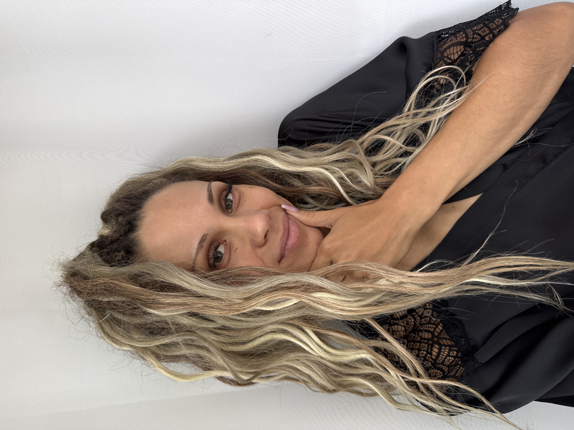 A woman with long, wavy blonde hair is pictured from the chest up, leaning against a white wall. She is wearing a black robe with lace trim on the sleeves and décolletage. Her right hand is placed on her chin, and she is looking towards the camera with a slight smile. The background is a plain white wall. There is no discernible urban environment or indication of Rusanovo, Russia.
