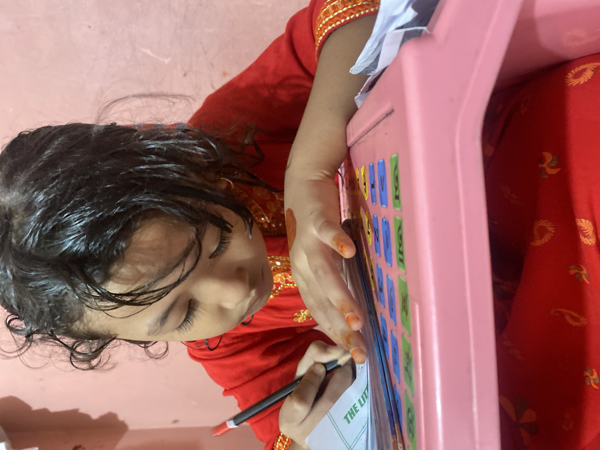 A young girl with dark, curly hair, wearing a red garment with gold embroidery, is seated at a pink desk, diligently writing with a pencil. Her gaze is focused downwards on a piece of paper with green lettering that reads "THE LI" visible, positioned over a colorful, textured surface that appears to be a learning board with letters and numbers. The girl's hands, adorned with orange henna, are actively engaged in the writing process. The background is a plain, pink wall, with a faint shadow cast on the left side. The lighting suggests an indoor setting, possibly a home environment in Dhaka, Bangladesh.