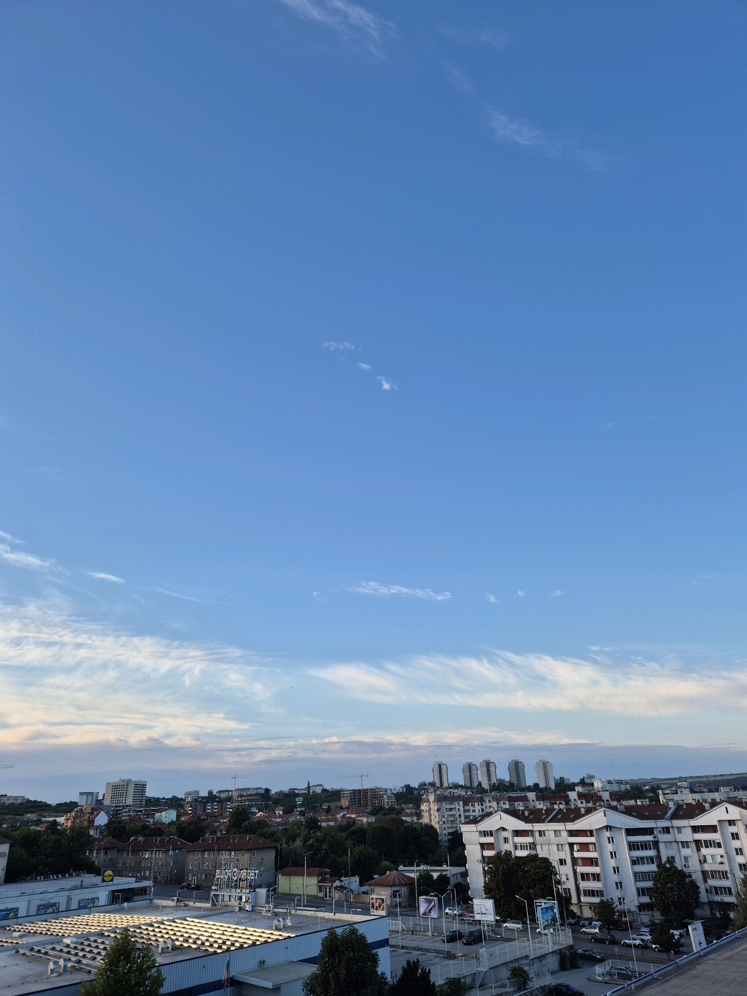 This image captures an urban landscape under a bright blue sky with wispy white clouds. In the foreground, a large, flat-roofed commercial building with numerous skylights is visible. Beyond this, a city skyline unfolds, featuring a mix of residential buildings, some with red-tiled roofs and white facades with red accents, interspersed with trees. Several taller, modern buildings and apartment complexes are visible in the distance, along with construction cranes, suggesting ongoing development. The scene appears to be a typical city view from an elevated vantage point during the daytime.