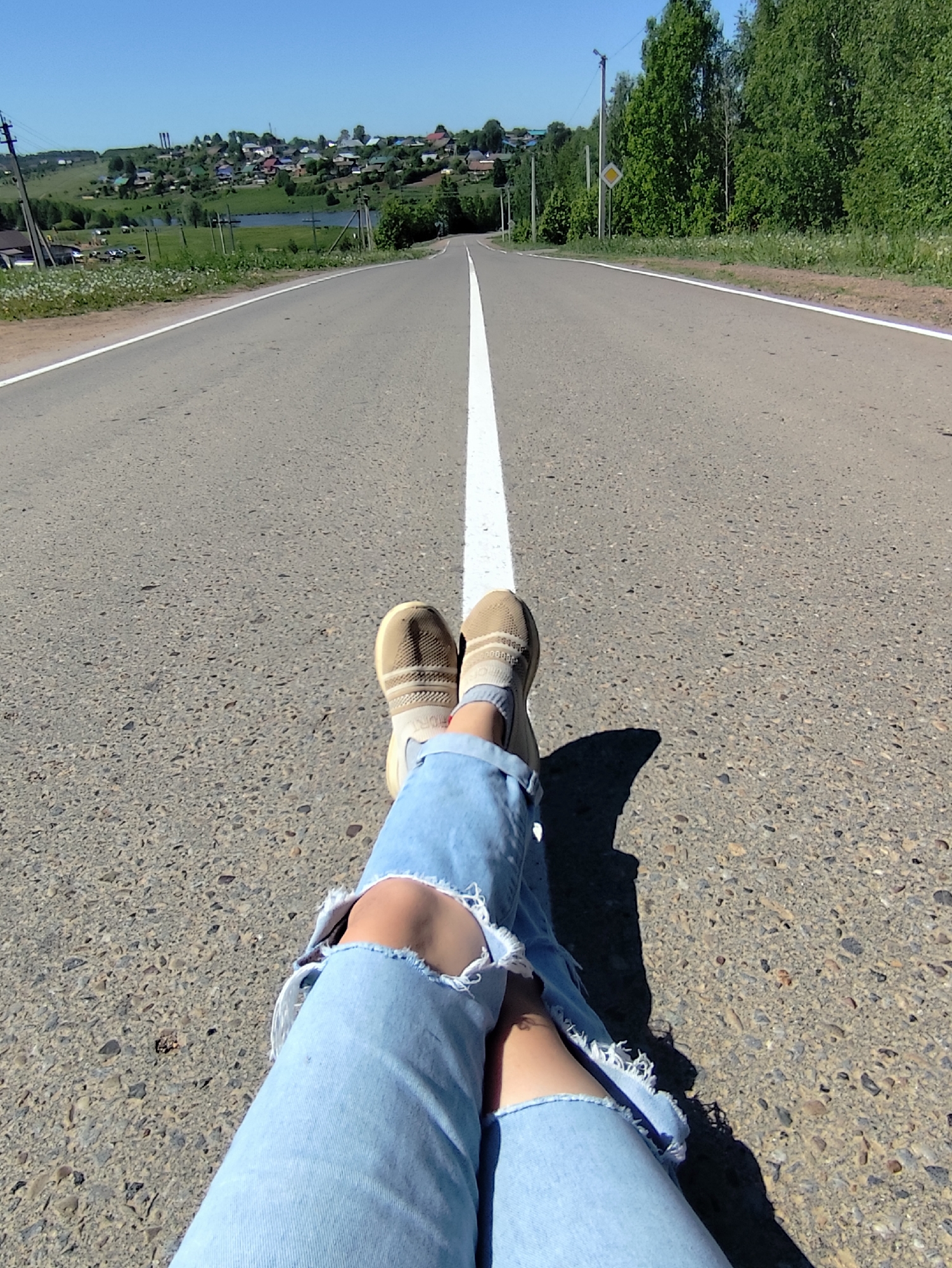 A straight asphalt road stretches into the distance, bisected by a solid white line. In the foreground, a person sits on the road with their legs extended towards the camera. They are wearing light blue ripped jeans and beige slip-on shoes with beige socks visible. Shadows cast by the person's legs are visible on the road surface. In the background, a village or small town is visible on a hill, with a body of water (likely a lake or pond) nearby. Trees and vegetation flank the sides of the road. A road sign is visible on the right side of the road. The image was taken in Фоки, Russia.