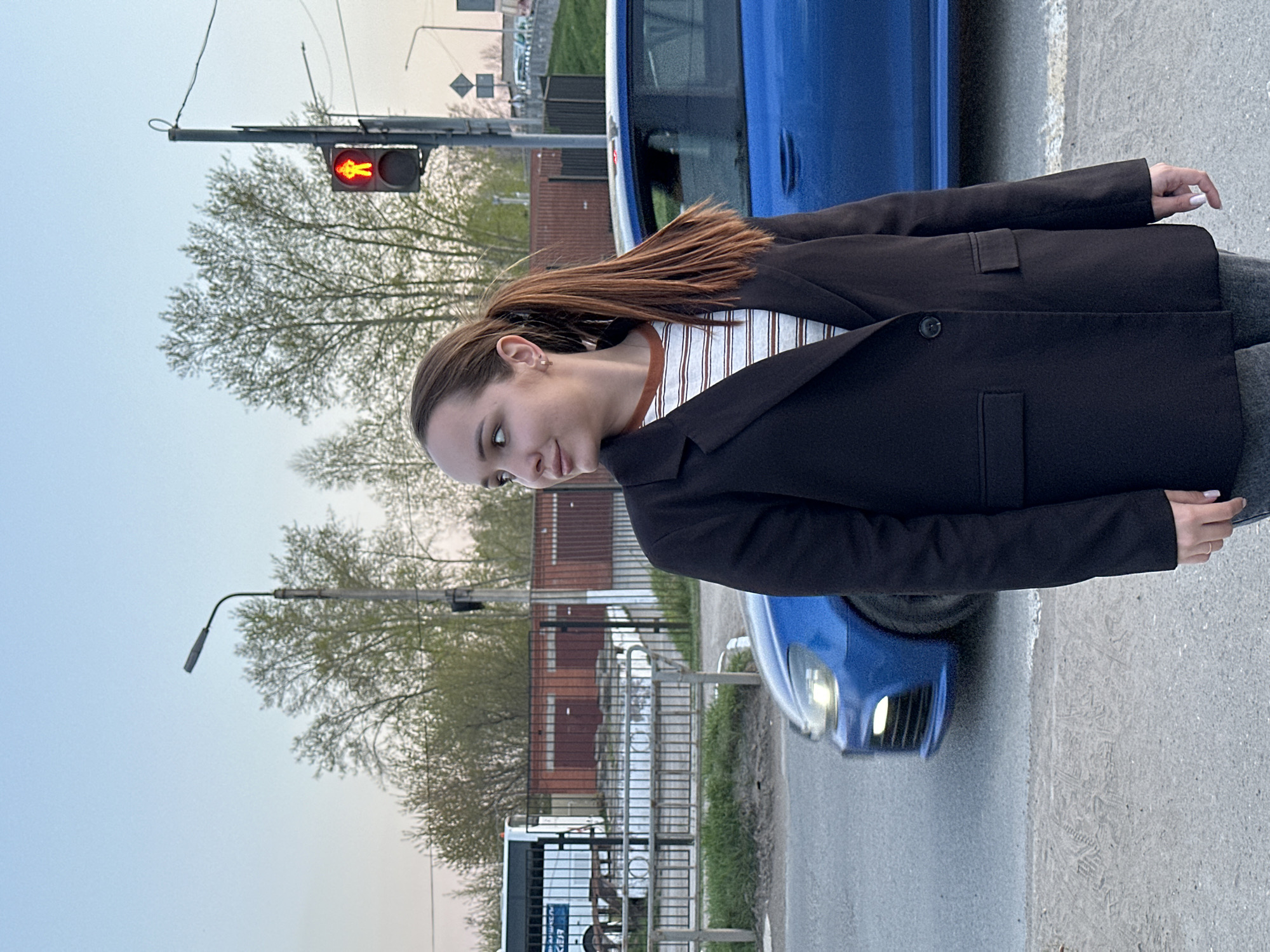 A woman stands on a crosswalk in Chaikovsky, Russia. She wears a dark blazer over a striped shirt and grey pants. A blue car is partially visible beside her. A pedestrian traffic light shows a red "do not walk" signal. A bus stop and streetlights are in the background. Trees with sparse foliage line the street. The sky is overcast. The ground is concrete or asphalt.